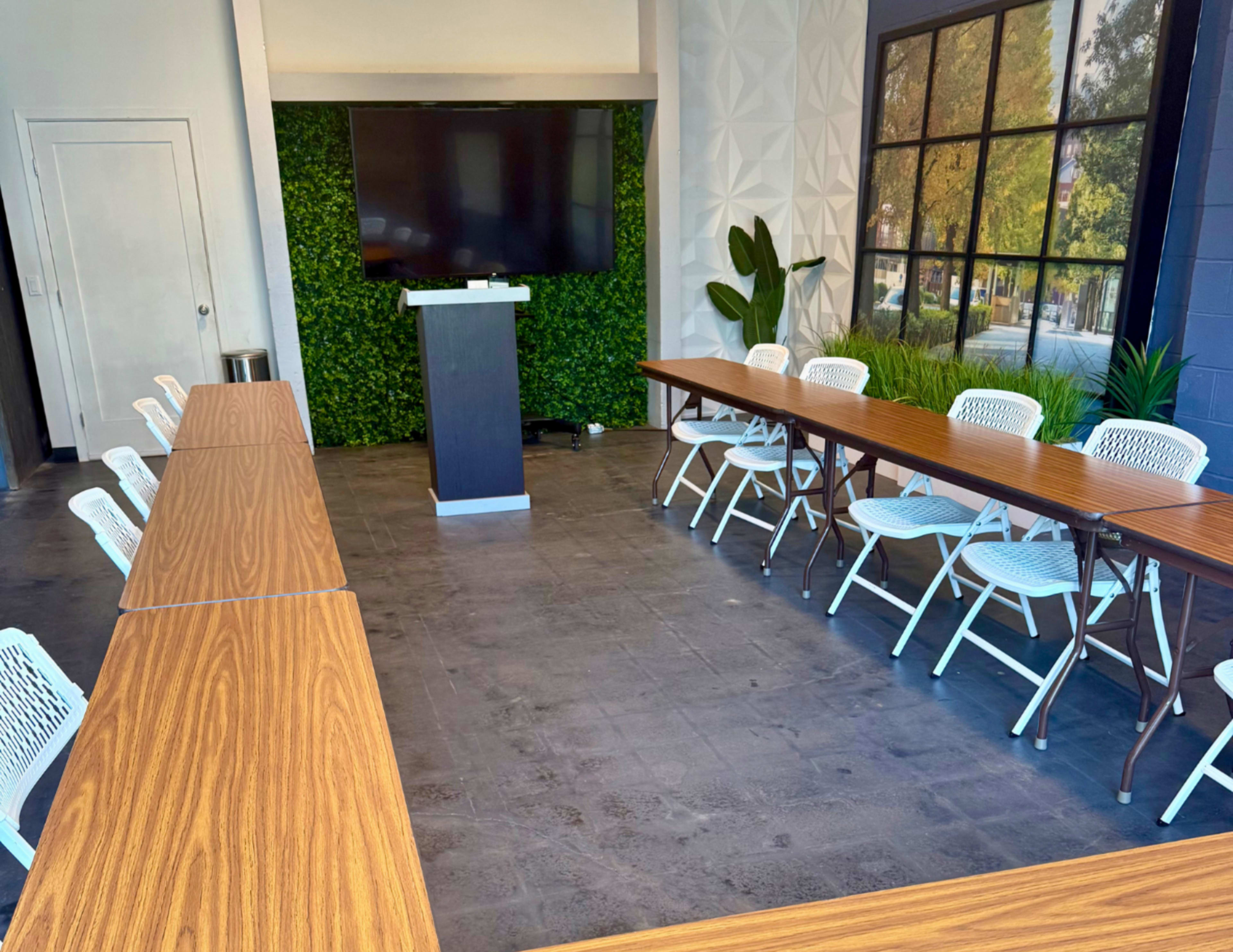The image shows a conference room with two long wooden tables arranged in an L-shape, surrounded by white chairs, and featuring a large screen mounted on a wall covered with greenery.
