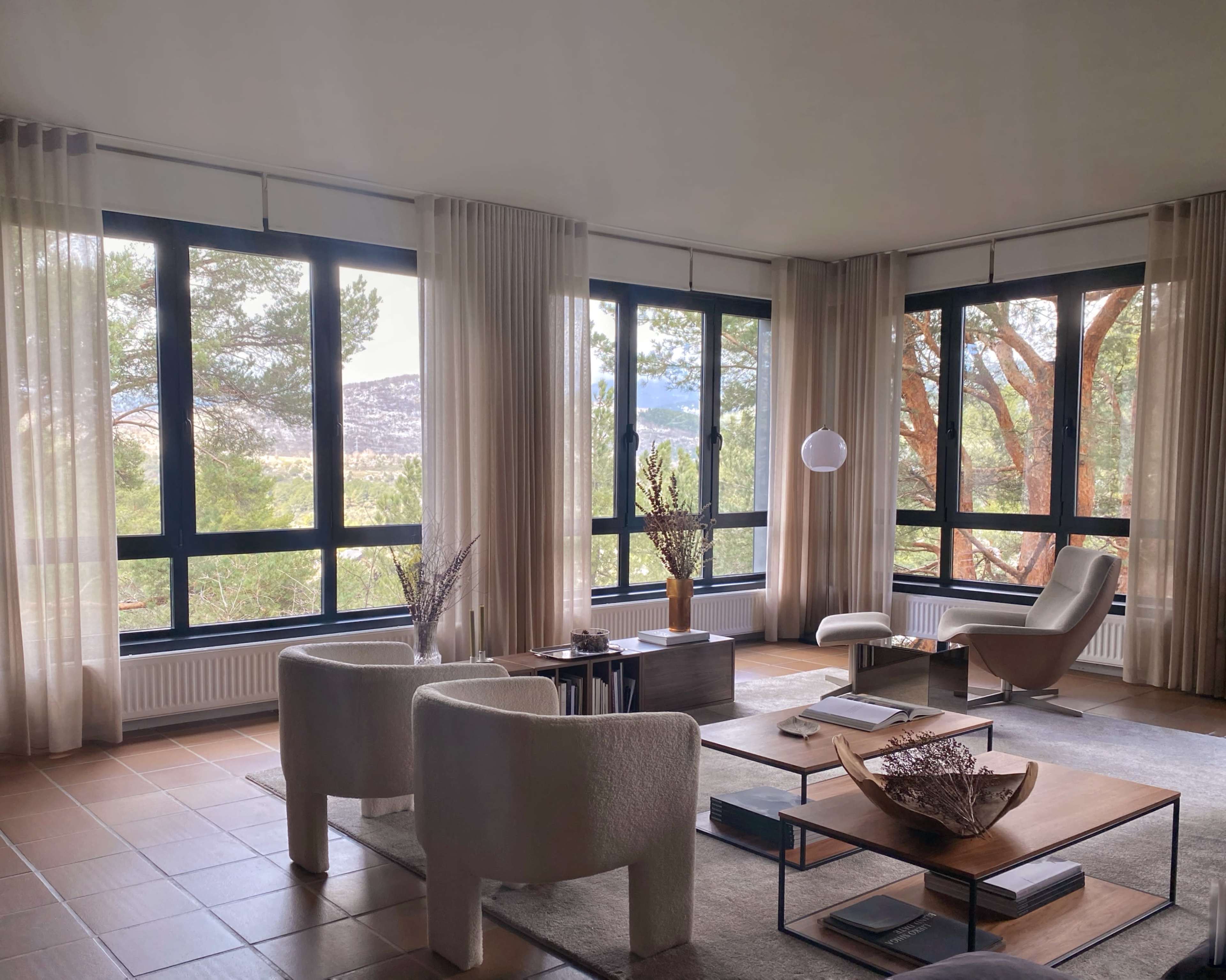 The image shows a modern living room with large windows, neutral-toned furniture, and a view of greenery outside.