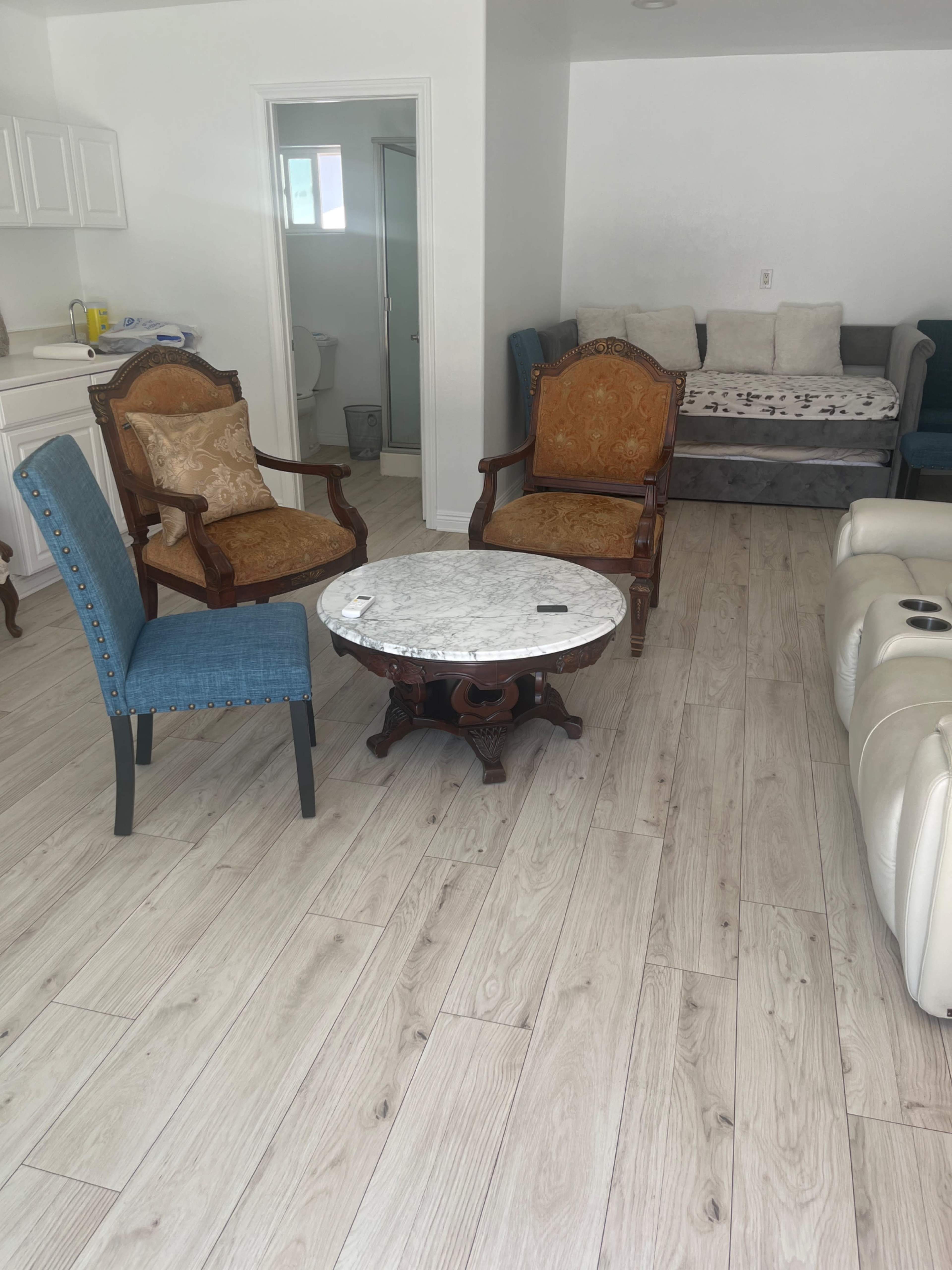 The image shows a living room with two ornate wooden chairs, a marble table, a blue upholstered chair, and a gray couch against a white wall.