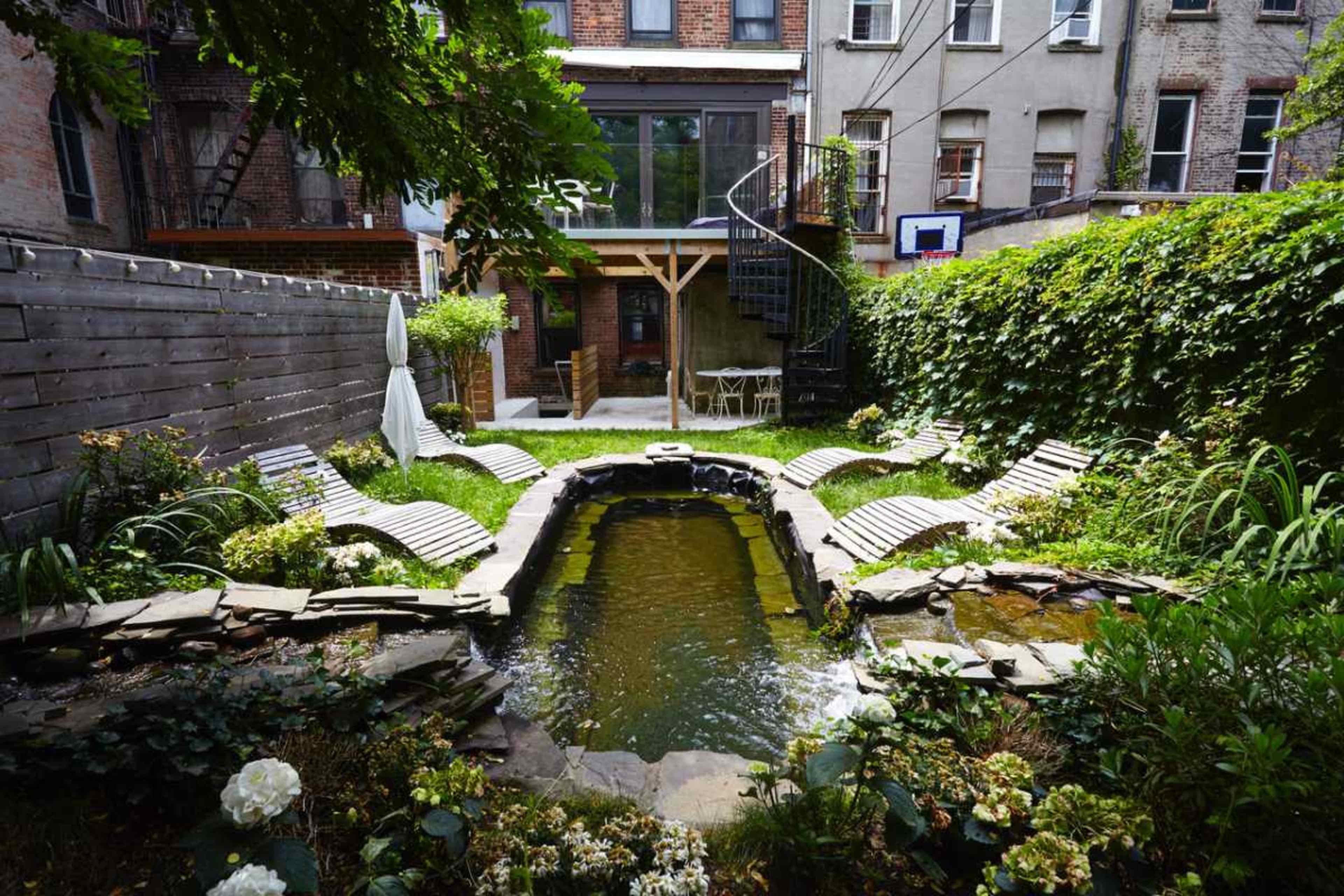 The image shows a garden with a small pond surrounded by stone paths and lounge chairs, framed by greenery and a spiral staircase leading to a building in the background.