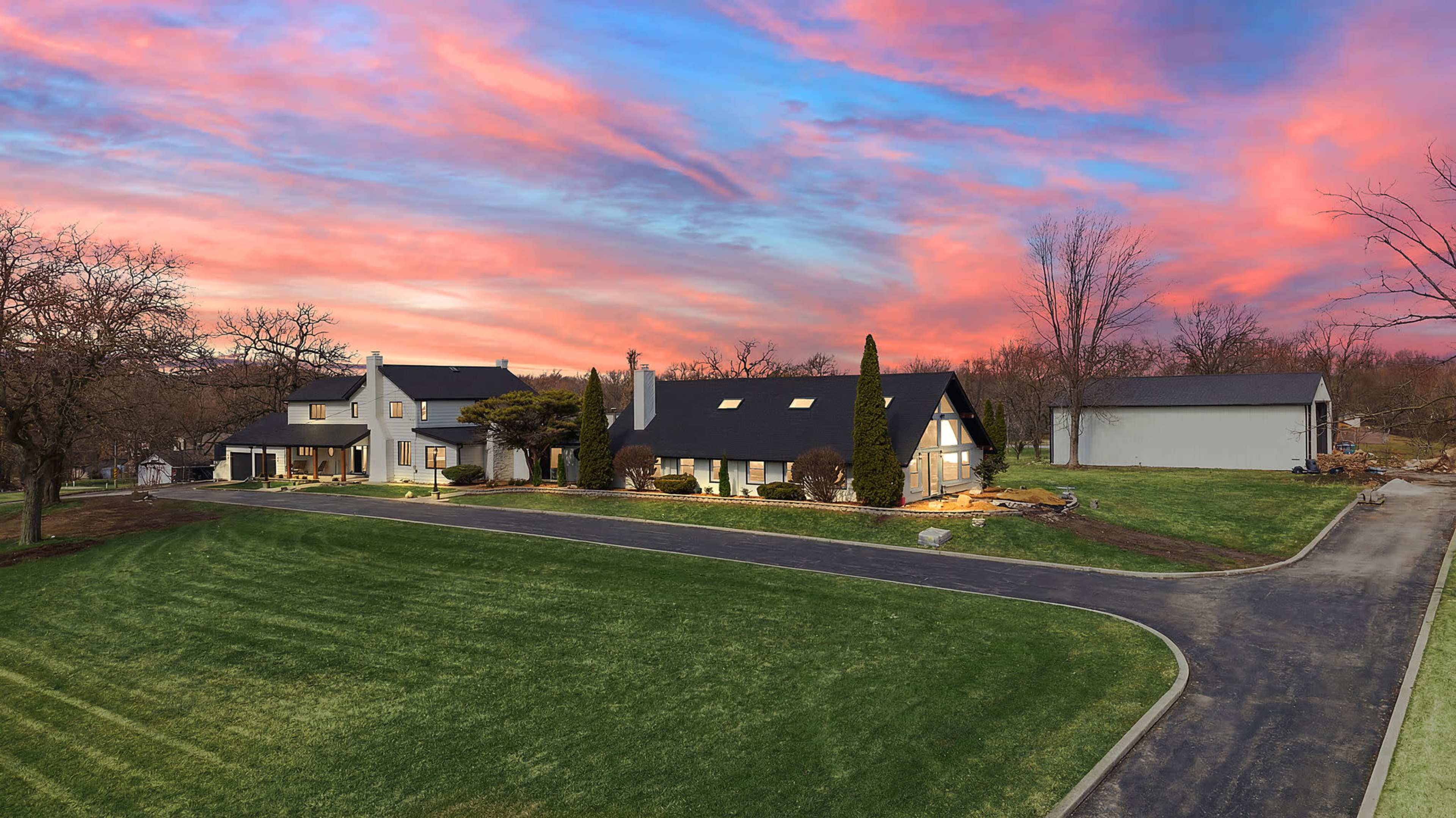 A large house with a dark roof is situated on a sprawling green lawn under a colorful sunset sky.