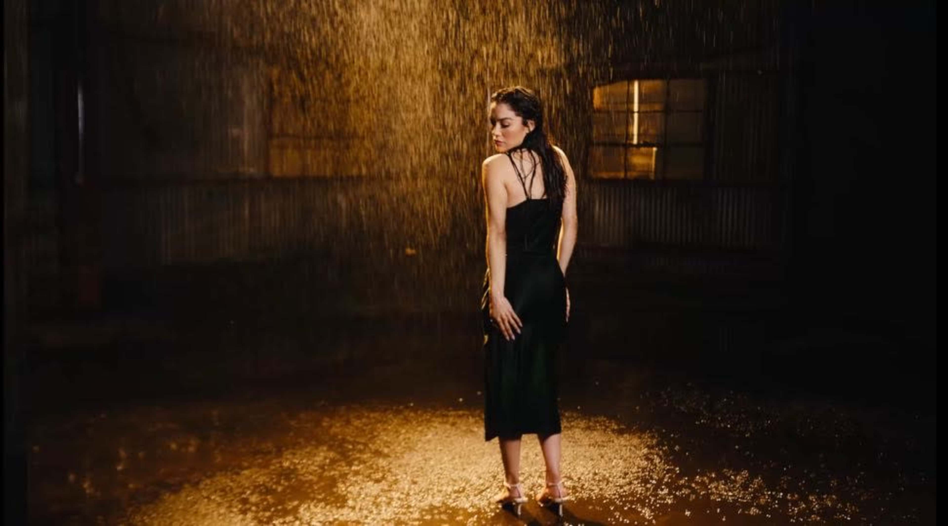 A woman in a black dress stands under a waterfall of rain in a dimly lit industrial space.
