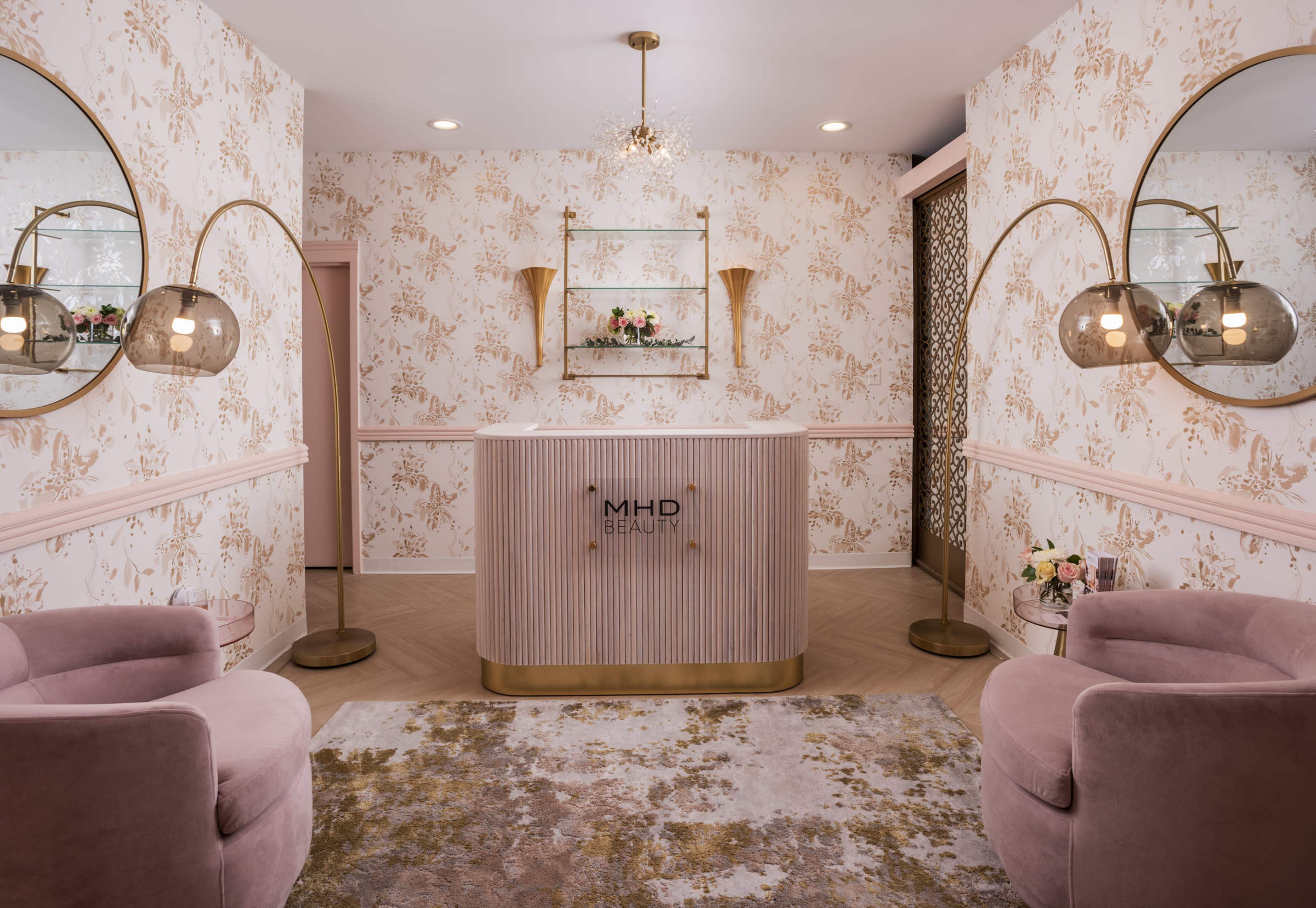 A modern beauty salon reception area with a curved front desk, pink chairs, decorative mirrors, and floral wallpaper.
