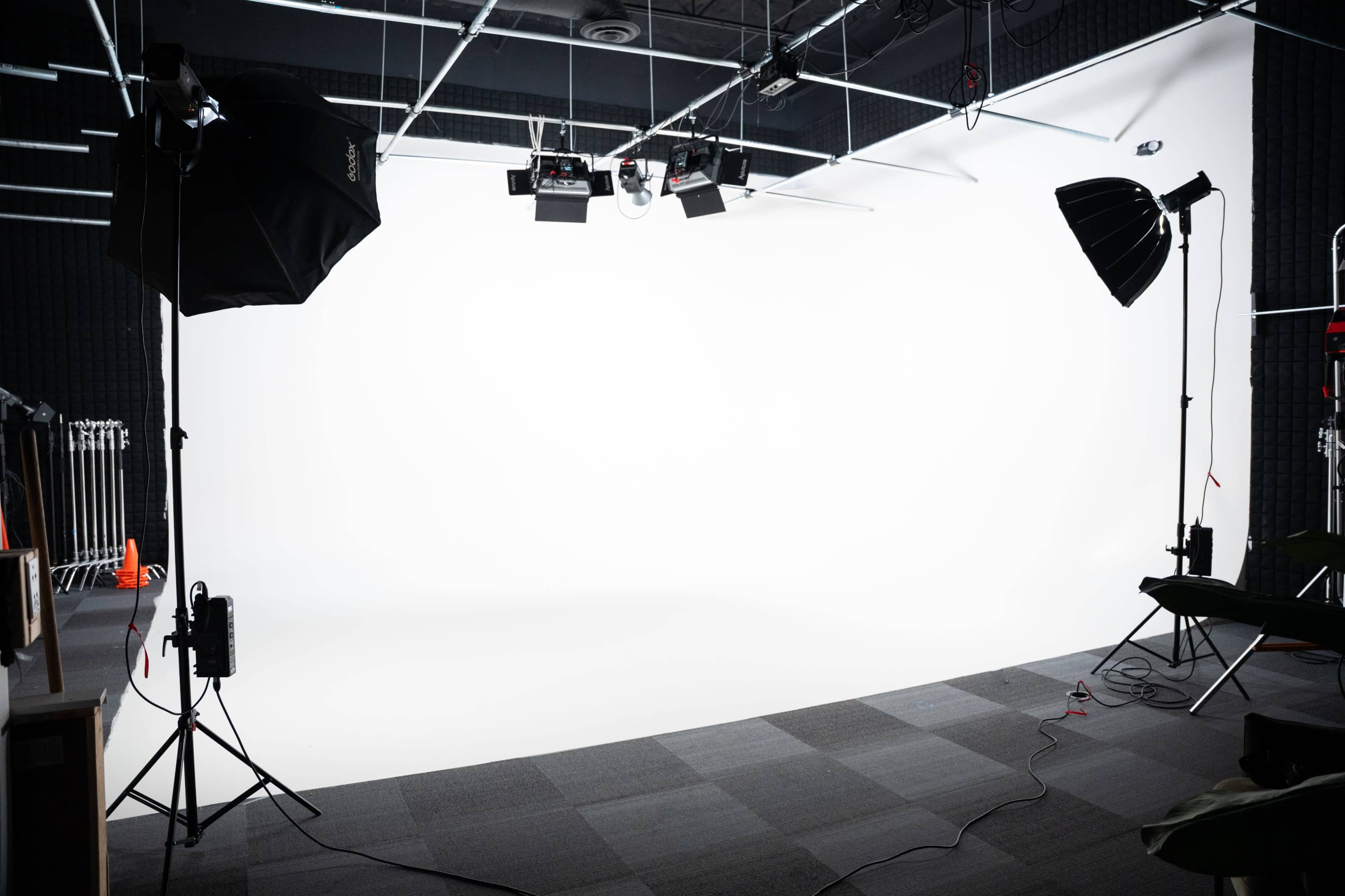 The image shows a photography studio with a blank white backdrop and various lighting equipment set up on either side.