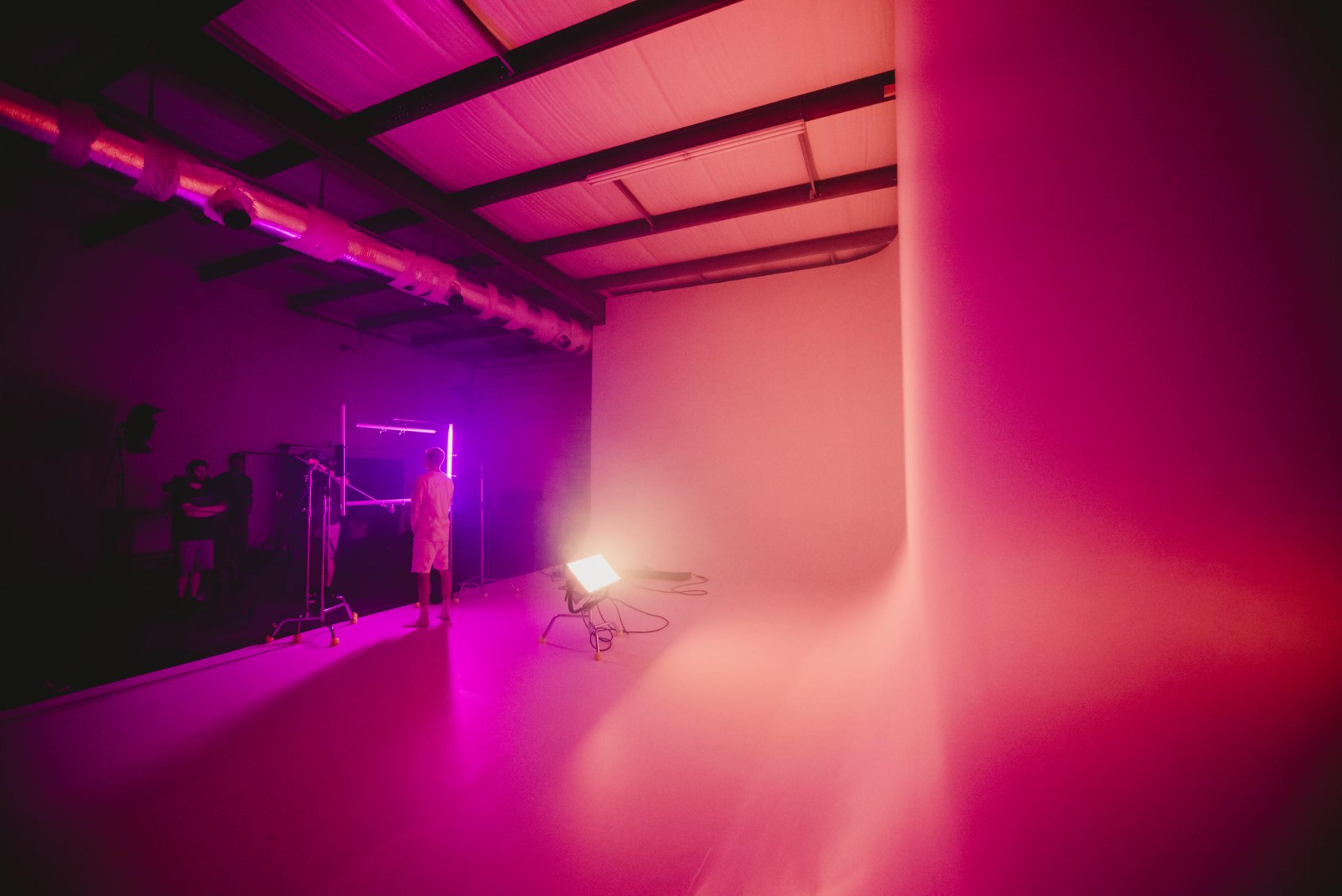 A person stands in a photography studio illuminated by pink and purple lights, with a seamless backdrop and equipment in the background.