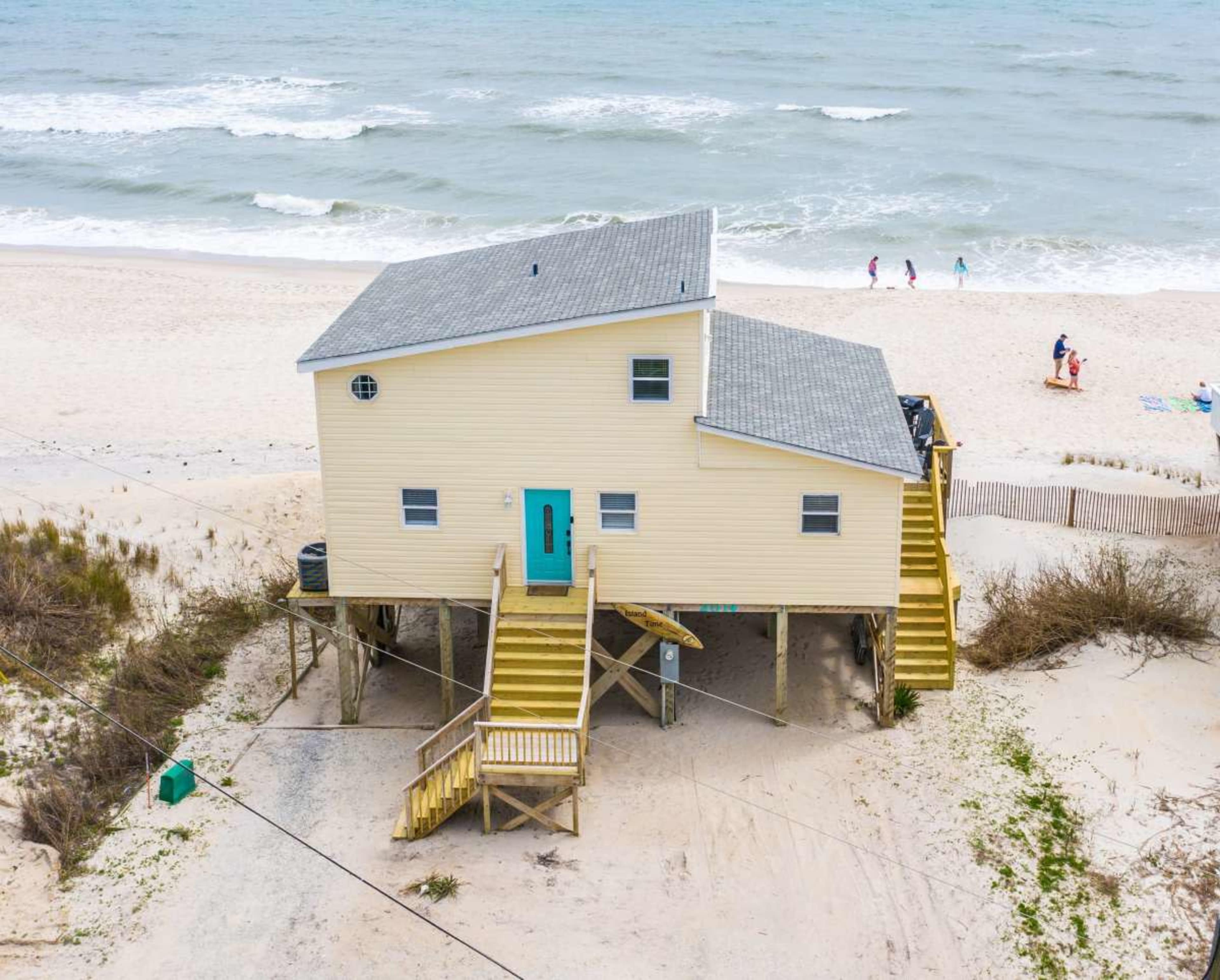 A yellow beach house on stilts is situated near a sandy shore, with waves in the background and people walking along the beach.