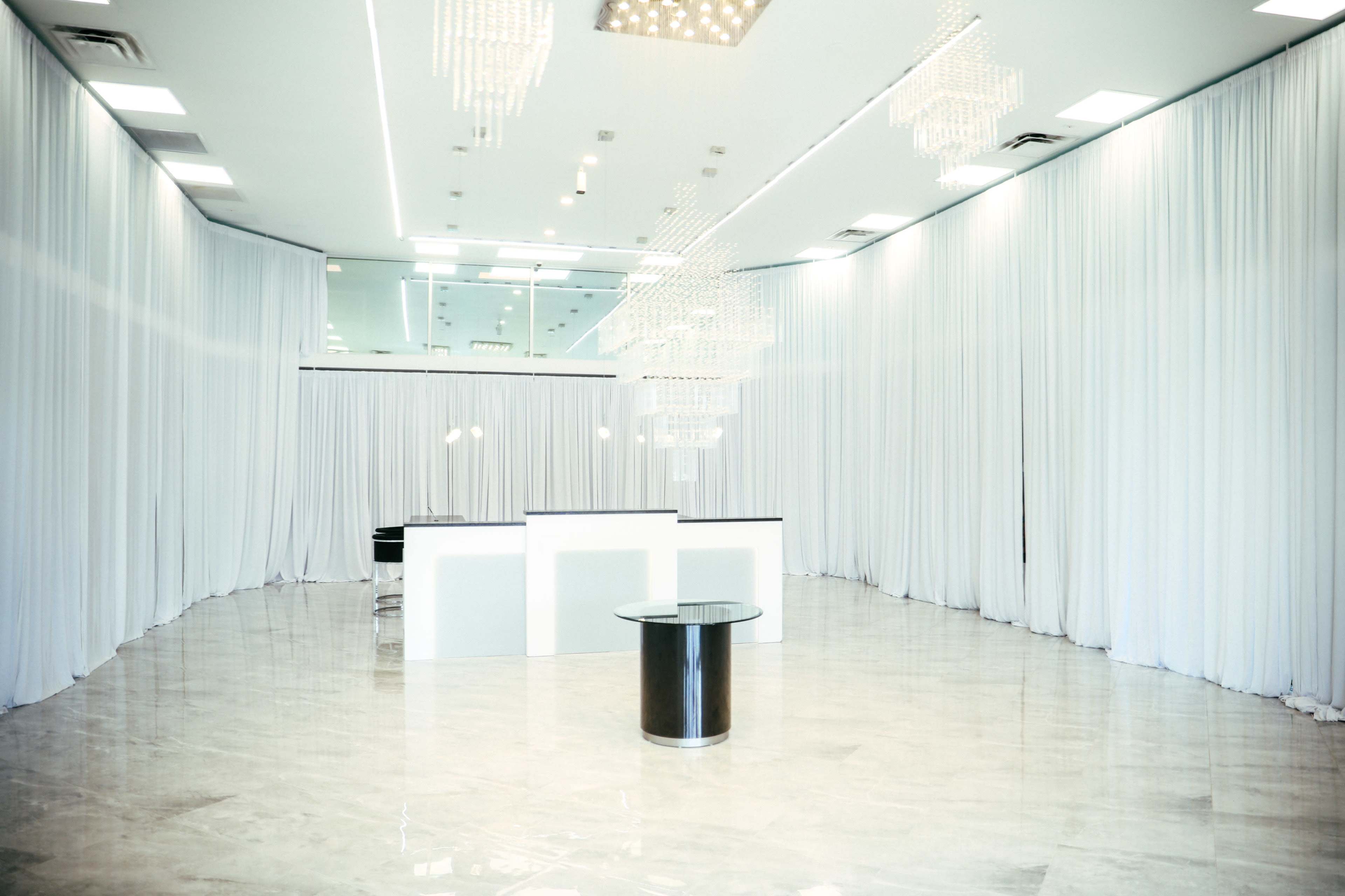 The image depicts a spacious interior with white draped curtains, polished floors, and a reception desk beneath hanging chandeliers.