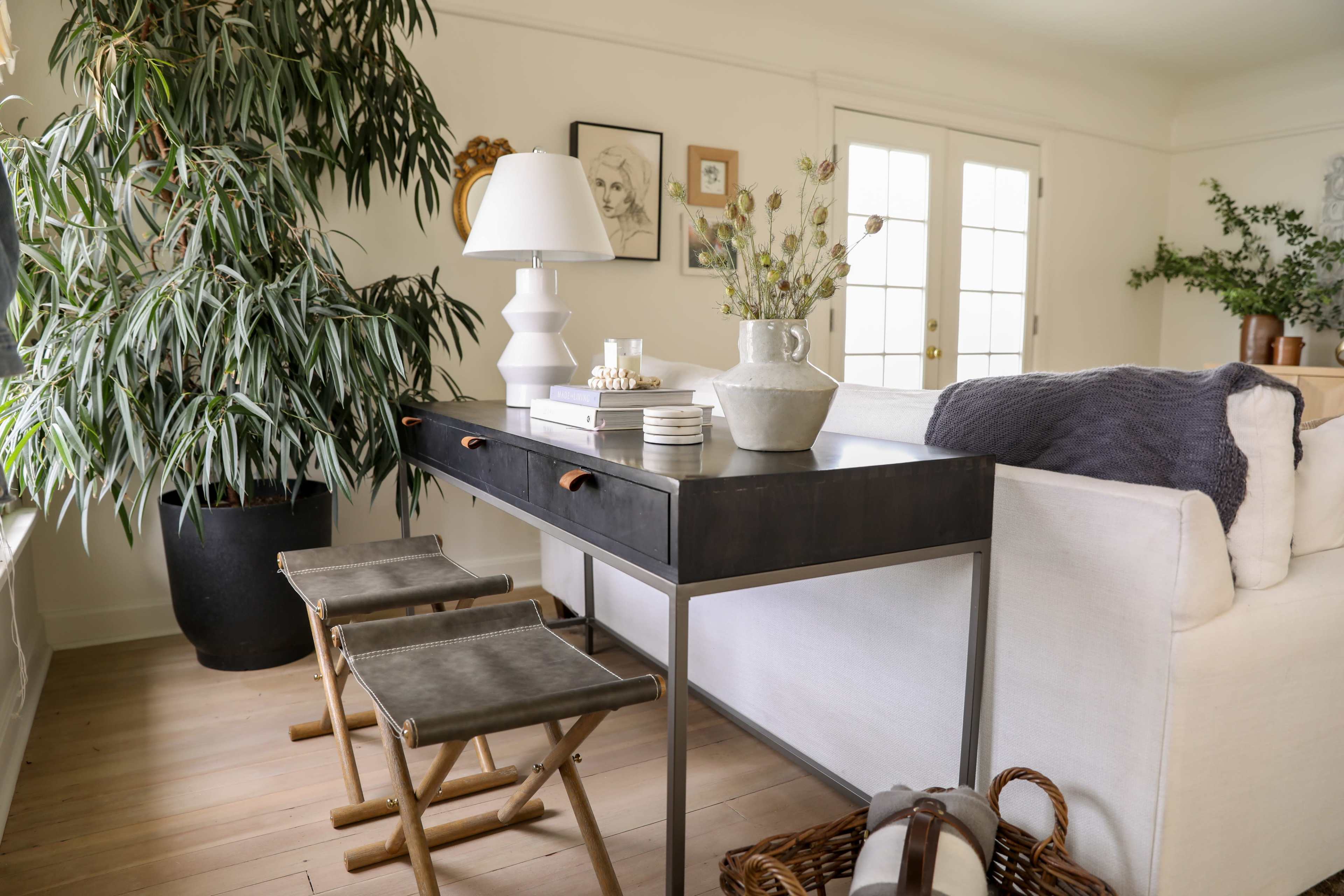 A modern interior space features a sleek black desk with decorative items, flanked by two wooden stools and a tall potted plant, against a backdrop of light walls and double doors.