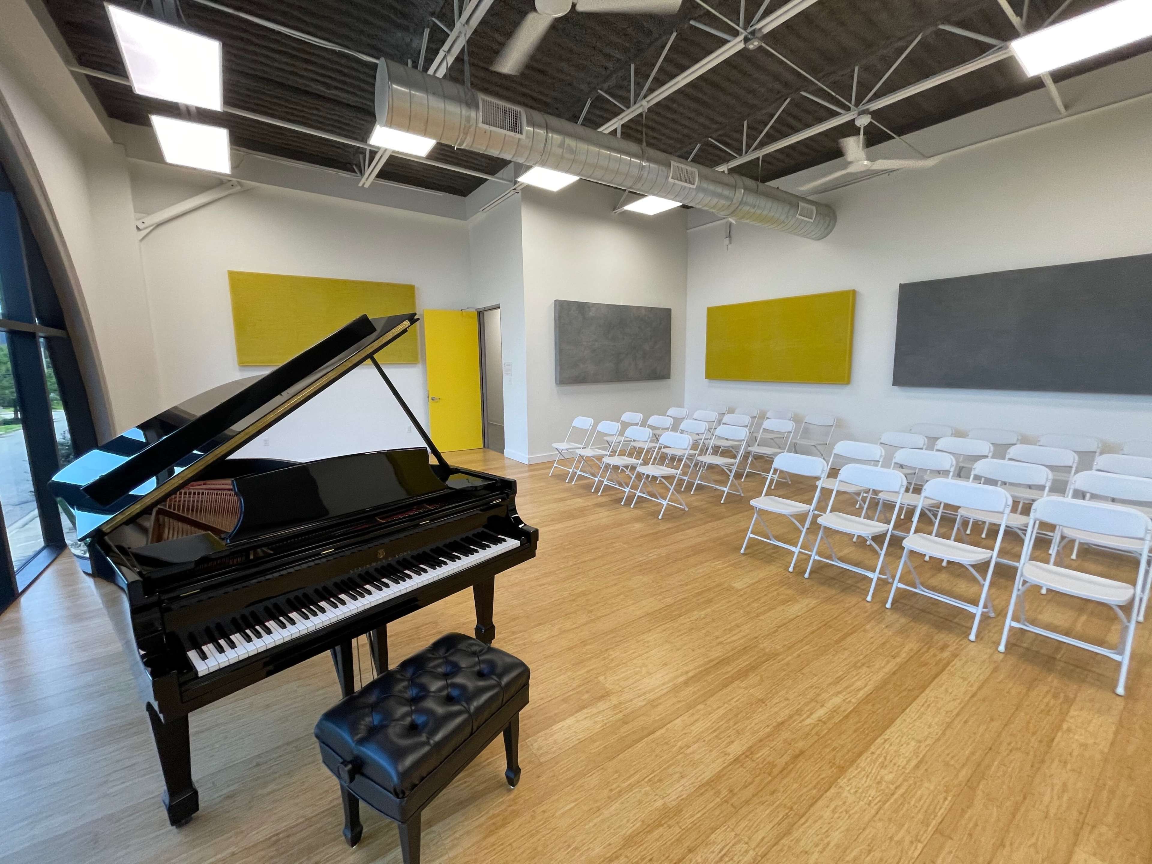 A black grand piano with a matching bench is positioned next to rows of white folding chairs in a well-lit room with yellow and gray wall panels.