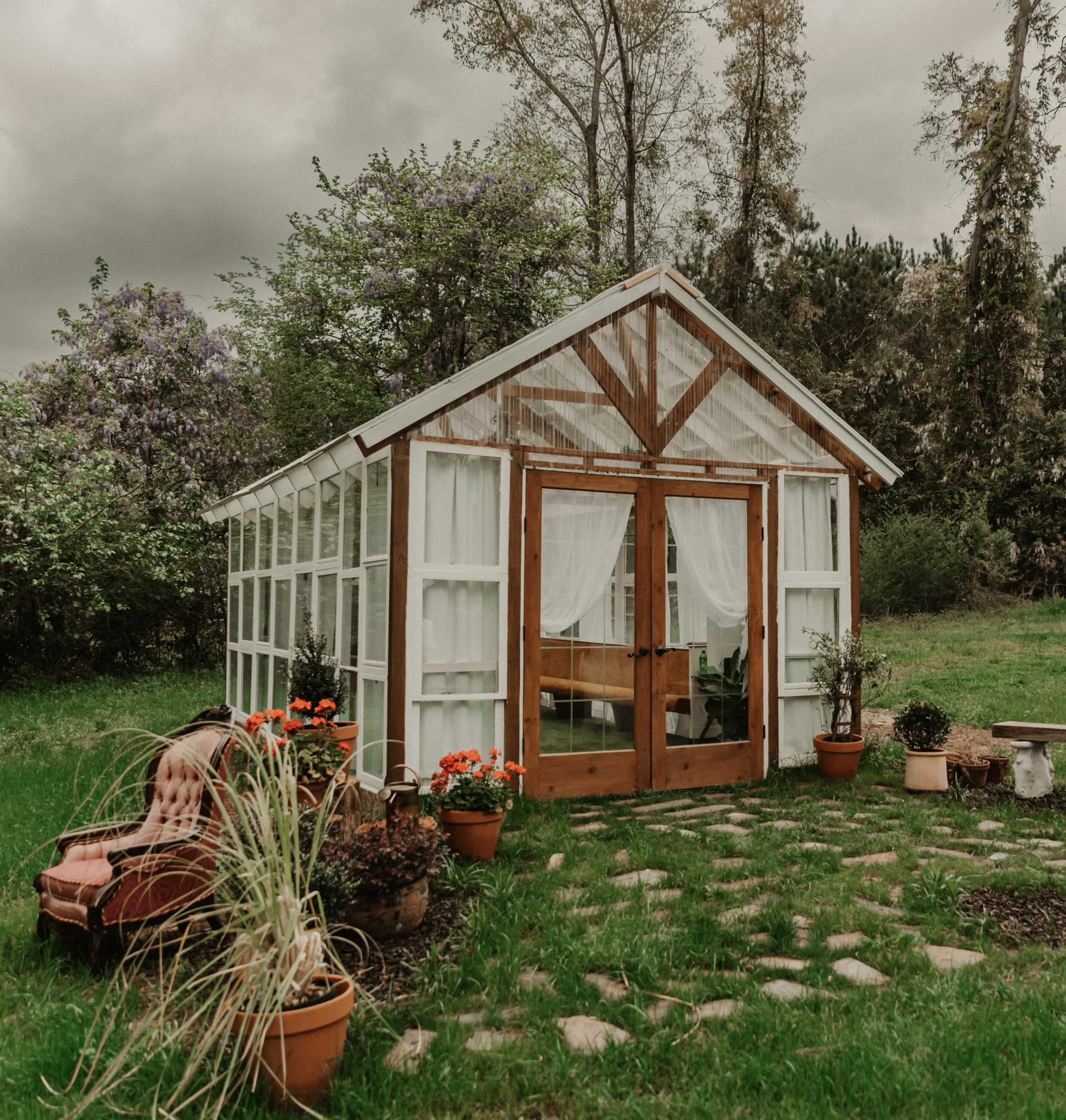 A white greenhouse with wooden doors and large windows is set on grassy land surrounded by flower pots and a vintage chair.