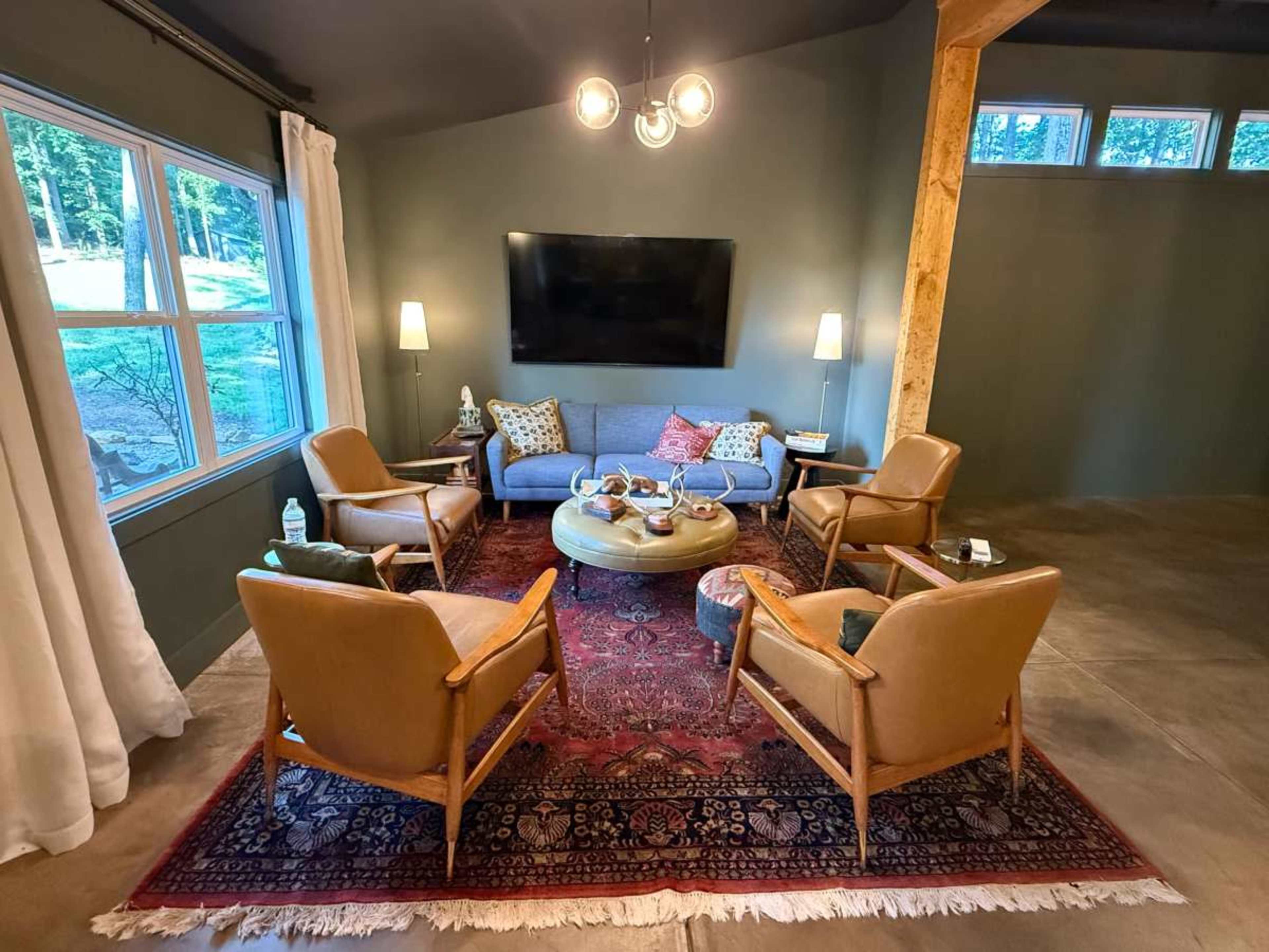 The image shows a cozy living room with a gray sofa, four brown chairs arranged around a round coffee table, and large windows letting in natural light.