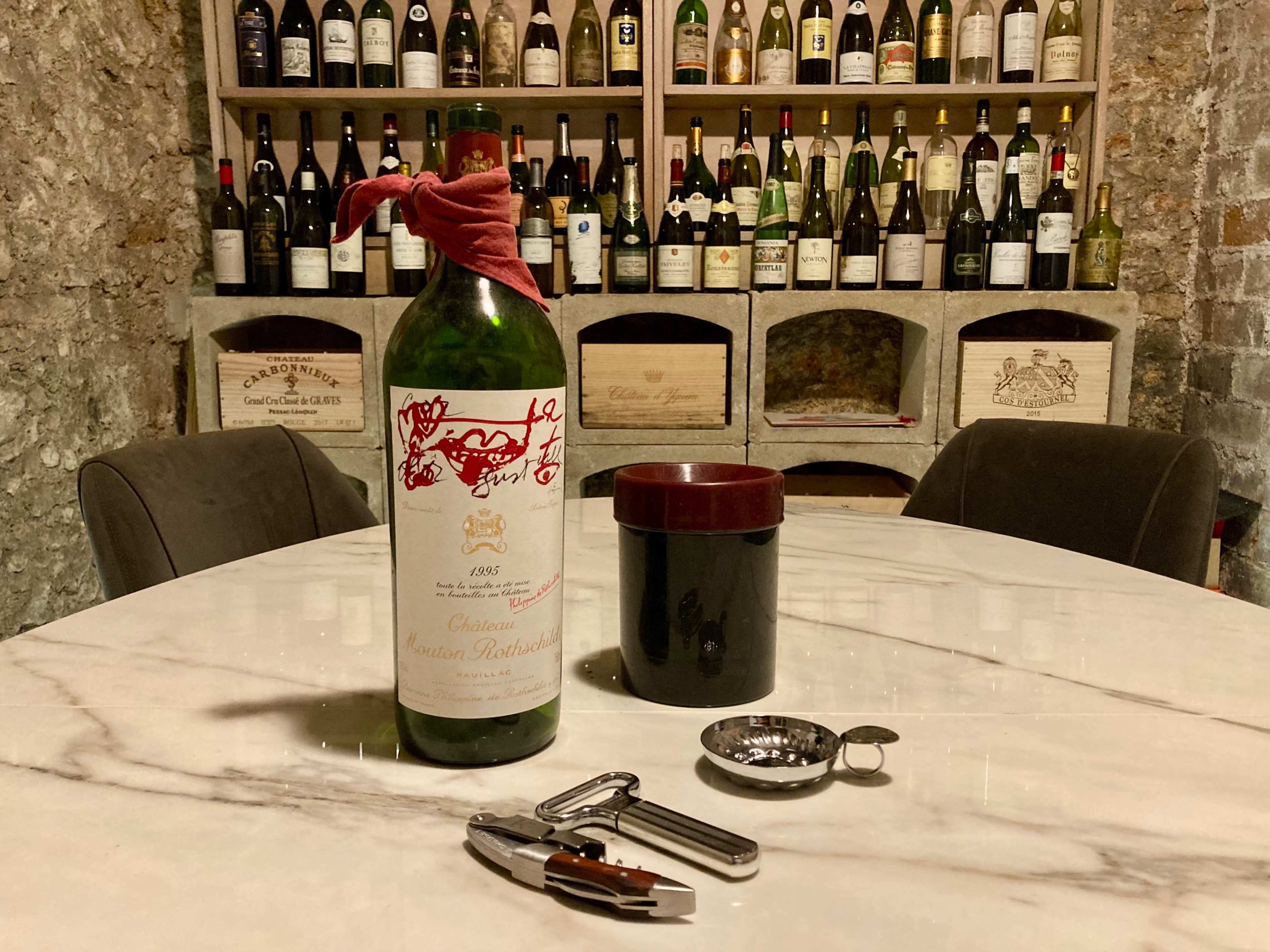 A striking wine bottle is displayed on a marble table along with a corkscrew, a wine aerator, and a glass, set against a backdrop of numerous wine bottles on shelves.