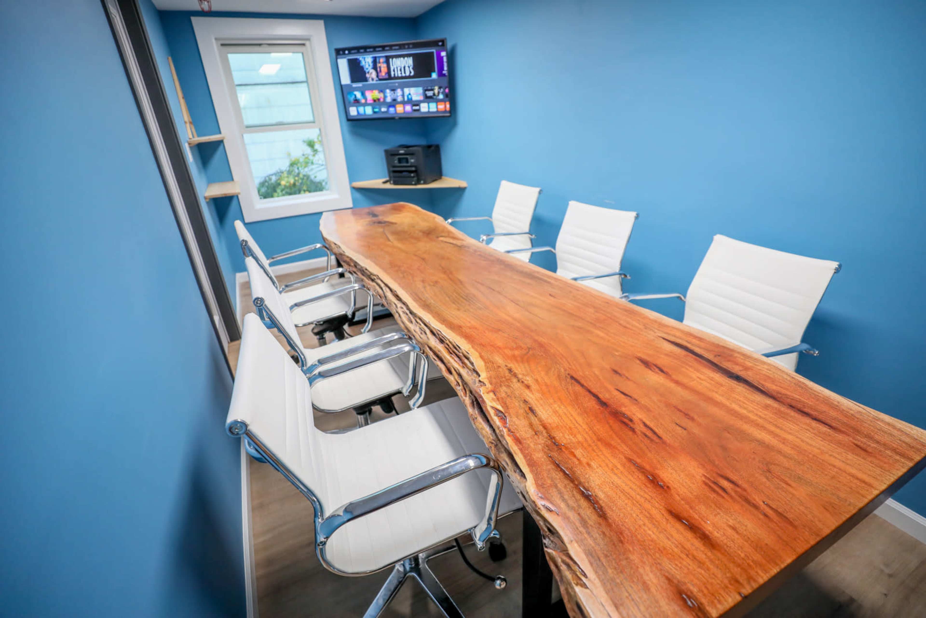 A long wooden conference table with sleek white chairs is placed in a room featuring blue walls and a TV mounted on the wall.