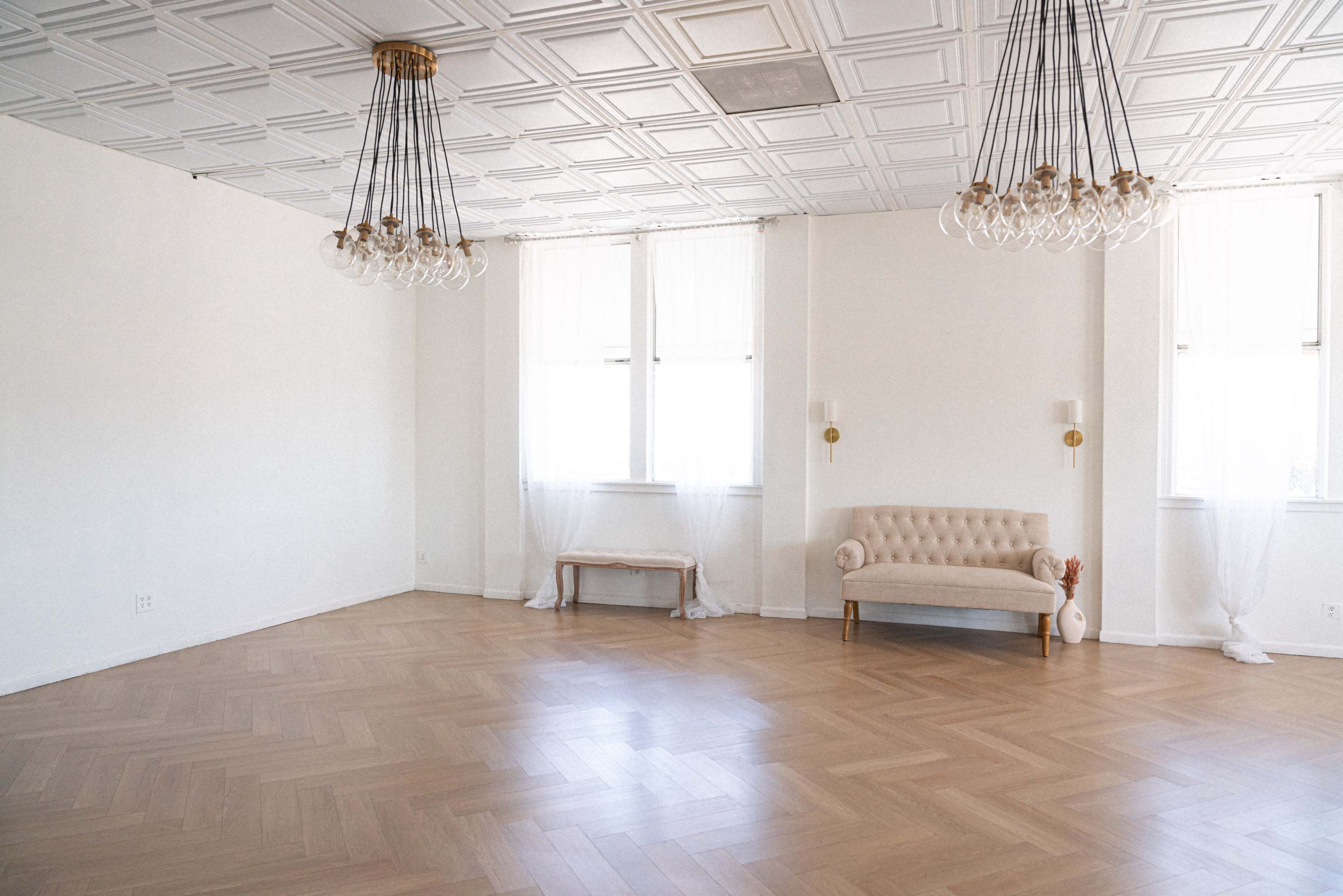 The image shows a spacious, well-lit room with wood flooring, large windows, and a tufted sofa against one wall.