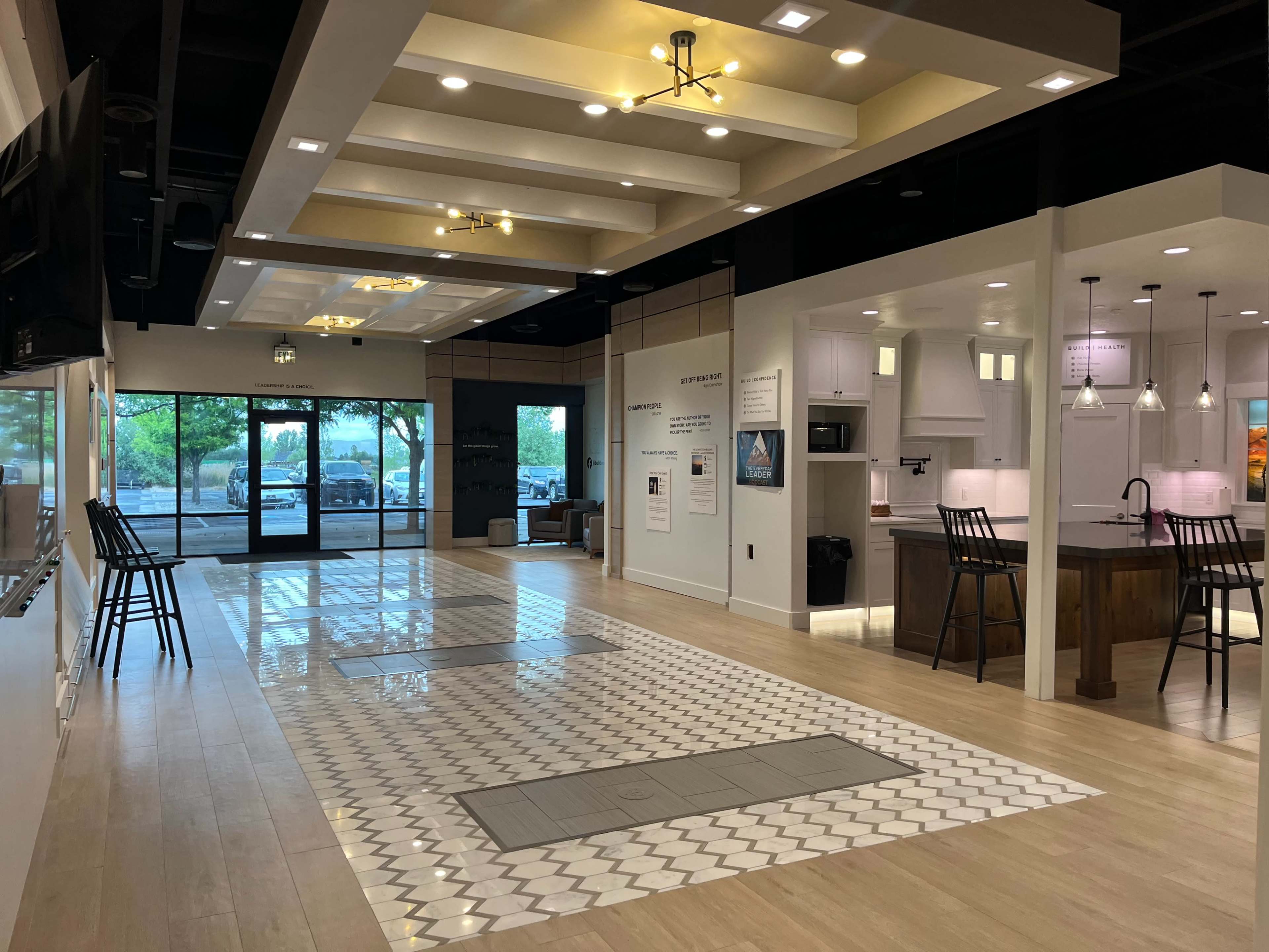 The image shows a modern, spacious interior featuring a kitchen area and seating, with large windows providing natural light and views of the exterior.