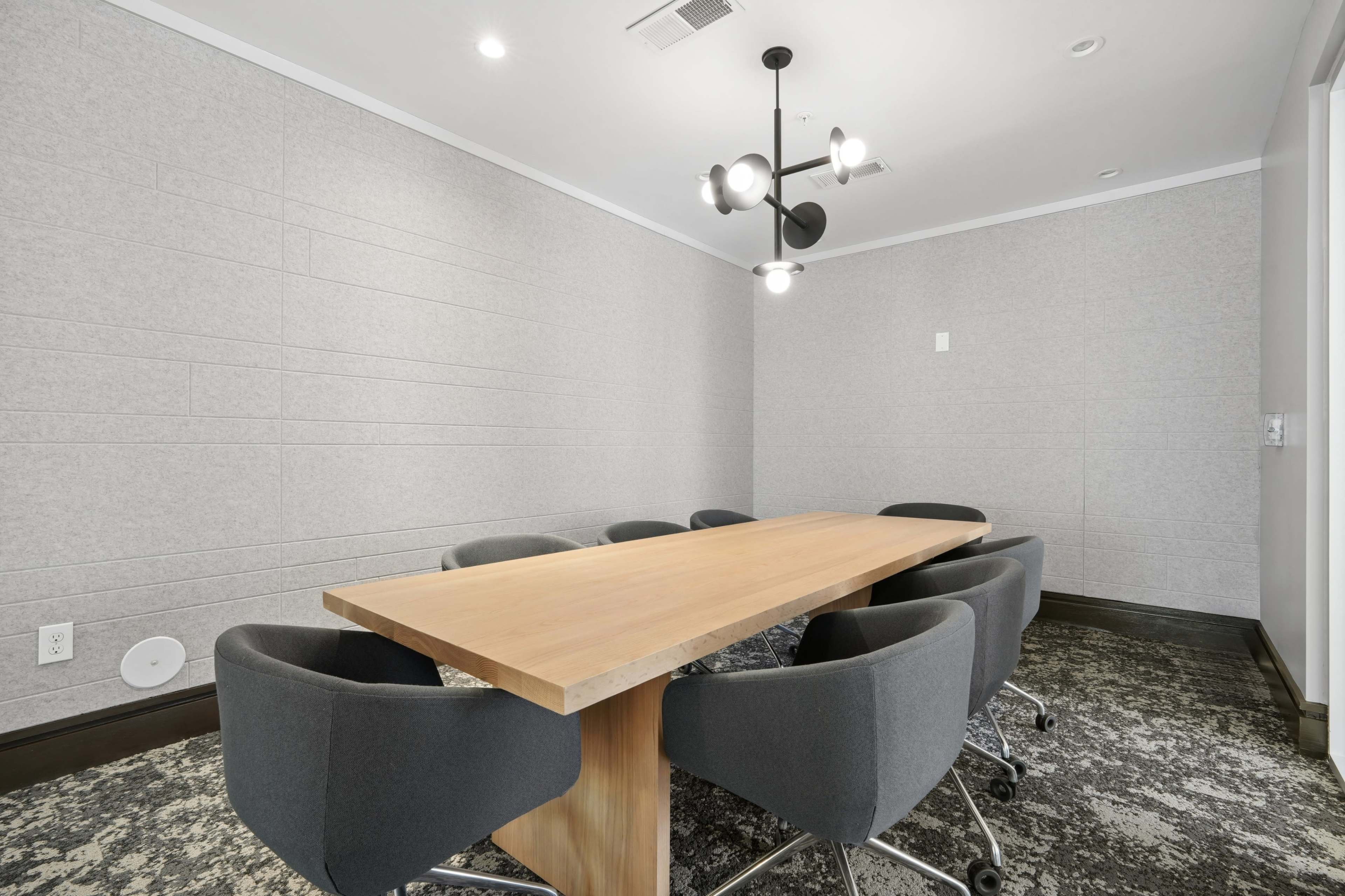 The image shows a modern meeting room with a long wooden table surrounded by gray swivel chairs and a contemporary ceiling light.