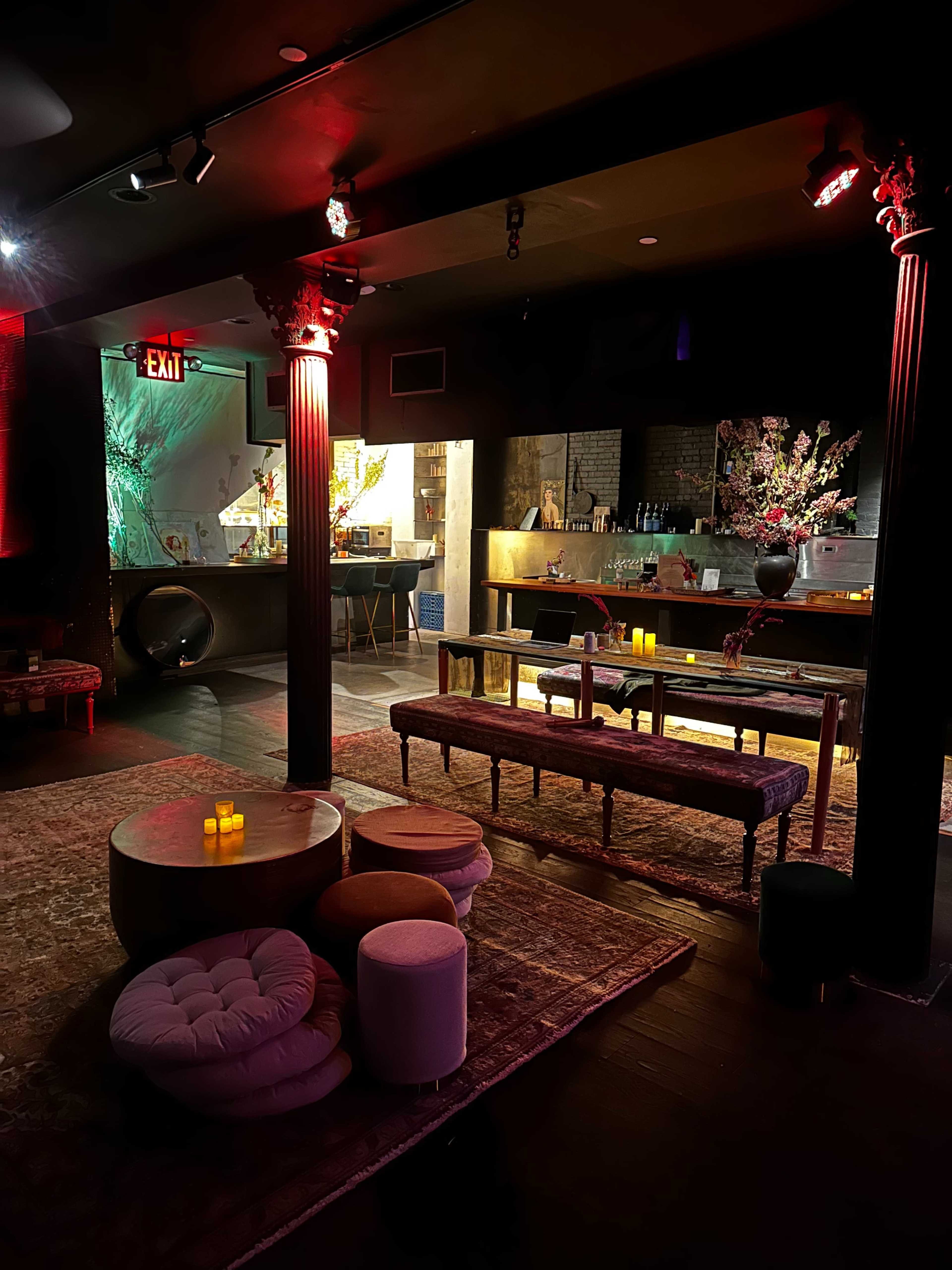 The image shows an interior of a dimly lit lounge with various seating options, a bar area in the background, and decorative elements including candles and floral arrangements.