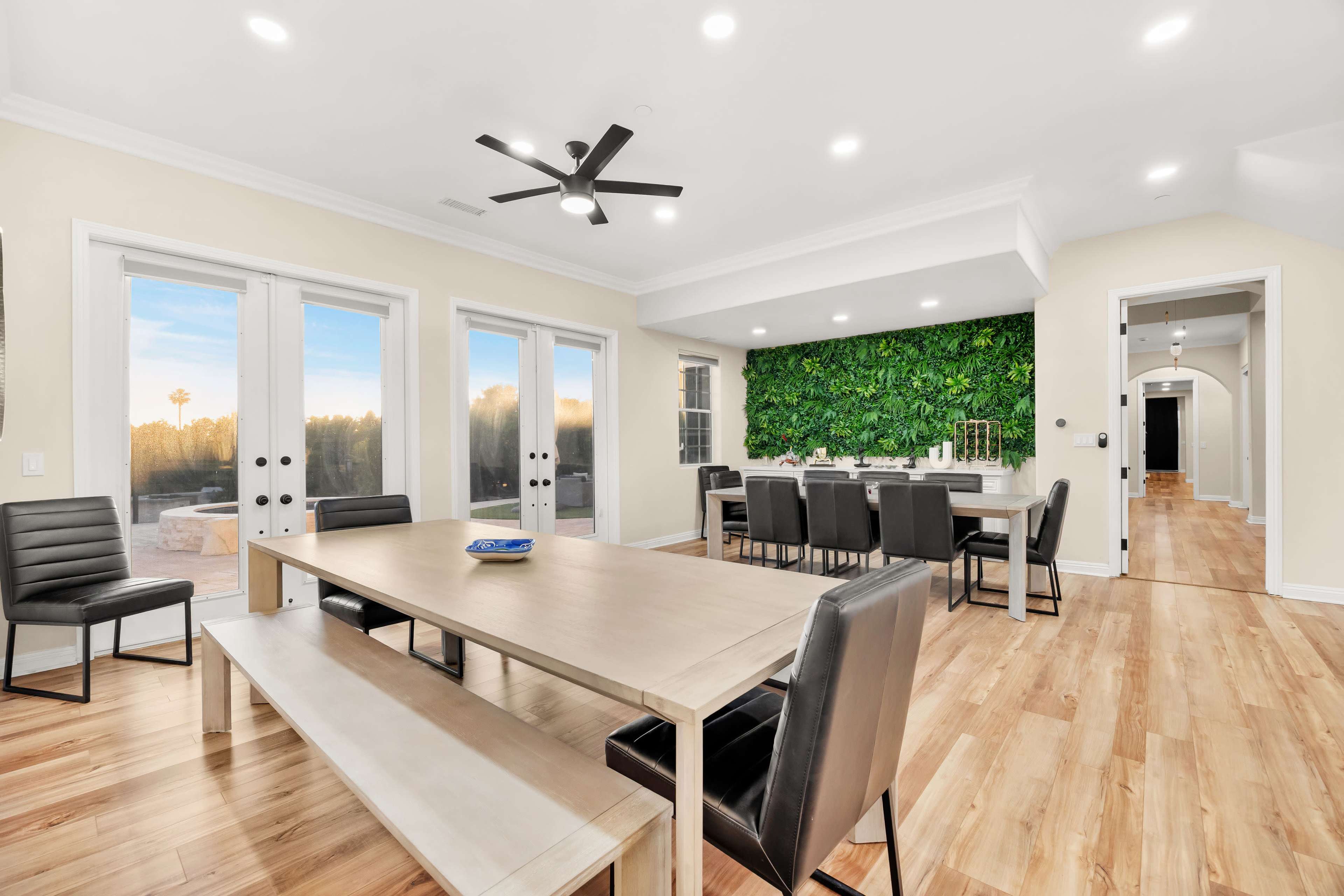 The image shows a modern dining area with a large wooden table, black chairs, and a green wall feature, illuminated by recessed lighting and large windows.