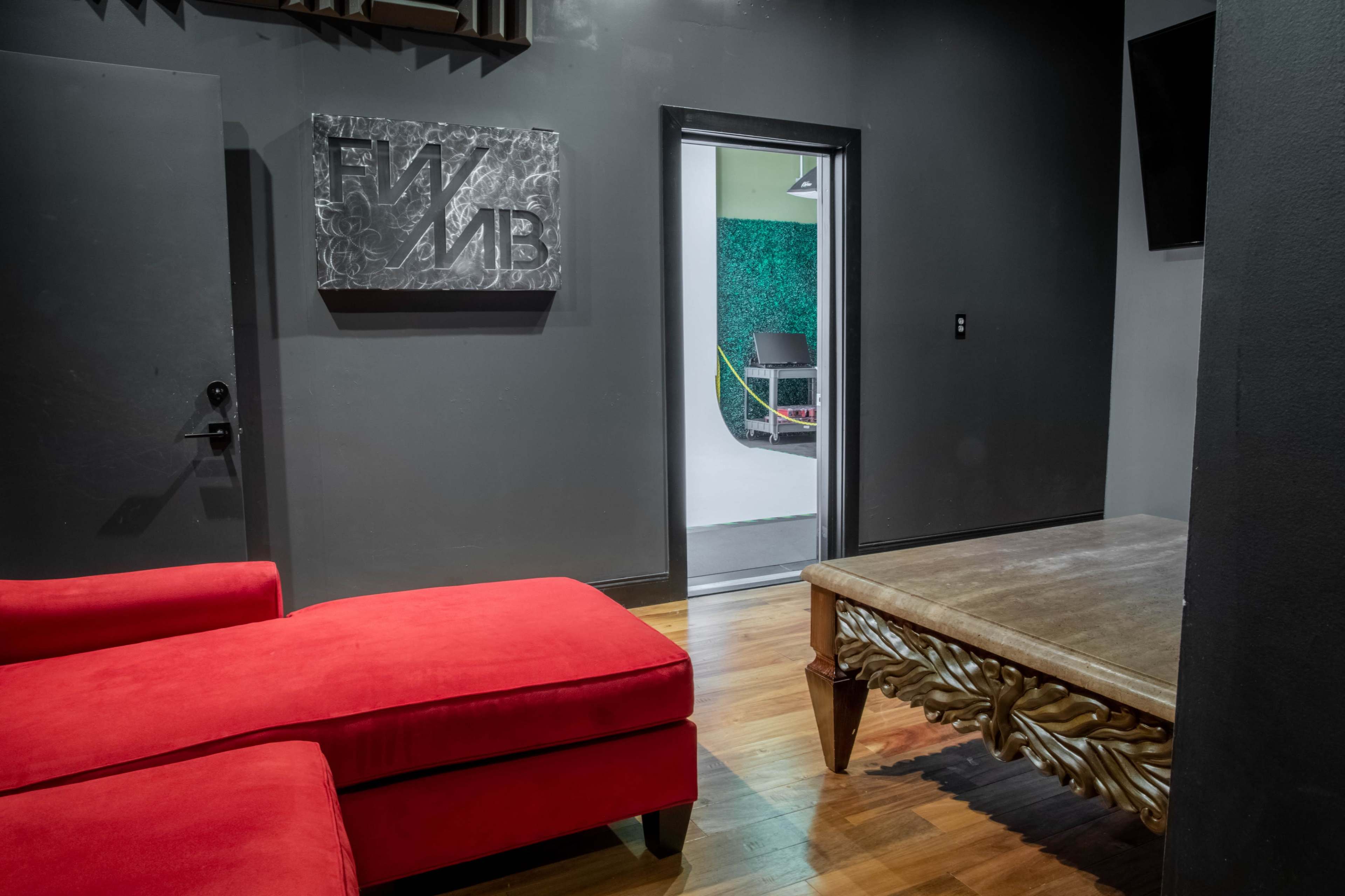 A red sectional sofa in a dimly lit room, with a wooden table and an open door leading to a green and white studio space beyond.