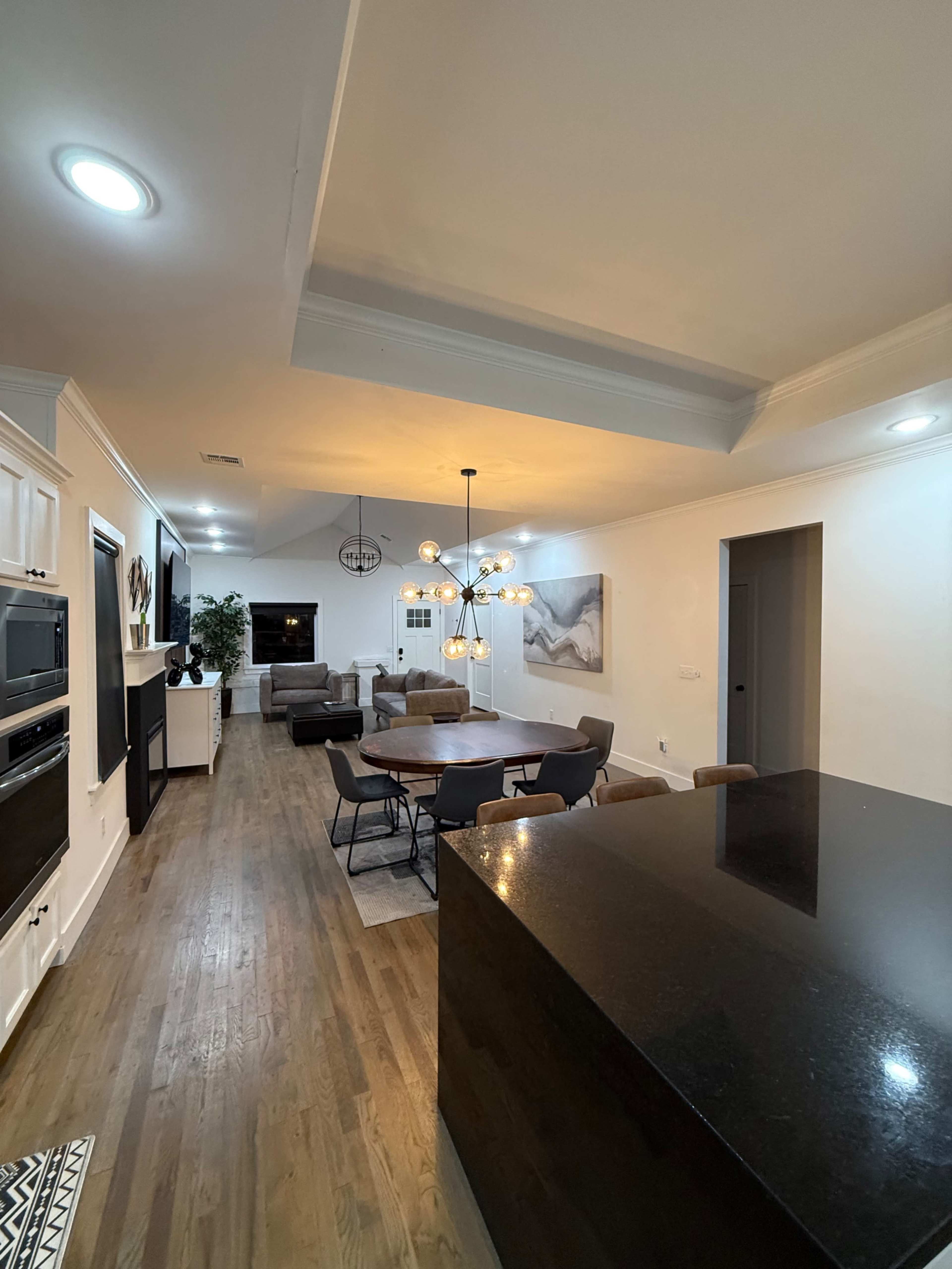 The image shows a modern kitchen and dining area with a dark countertop, wooden flooring, and a circular dining table surrounded by chairs under a decorative light fixture.