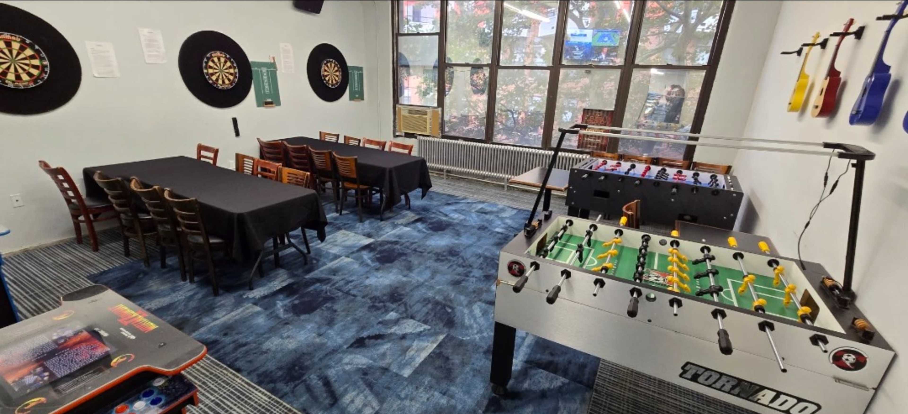 The room features a foosball table, dartboard, and seating arrangements, with large windows providing natural light.