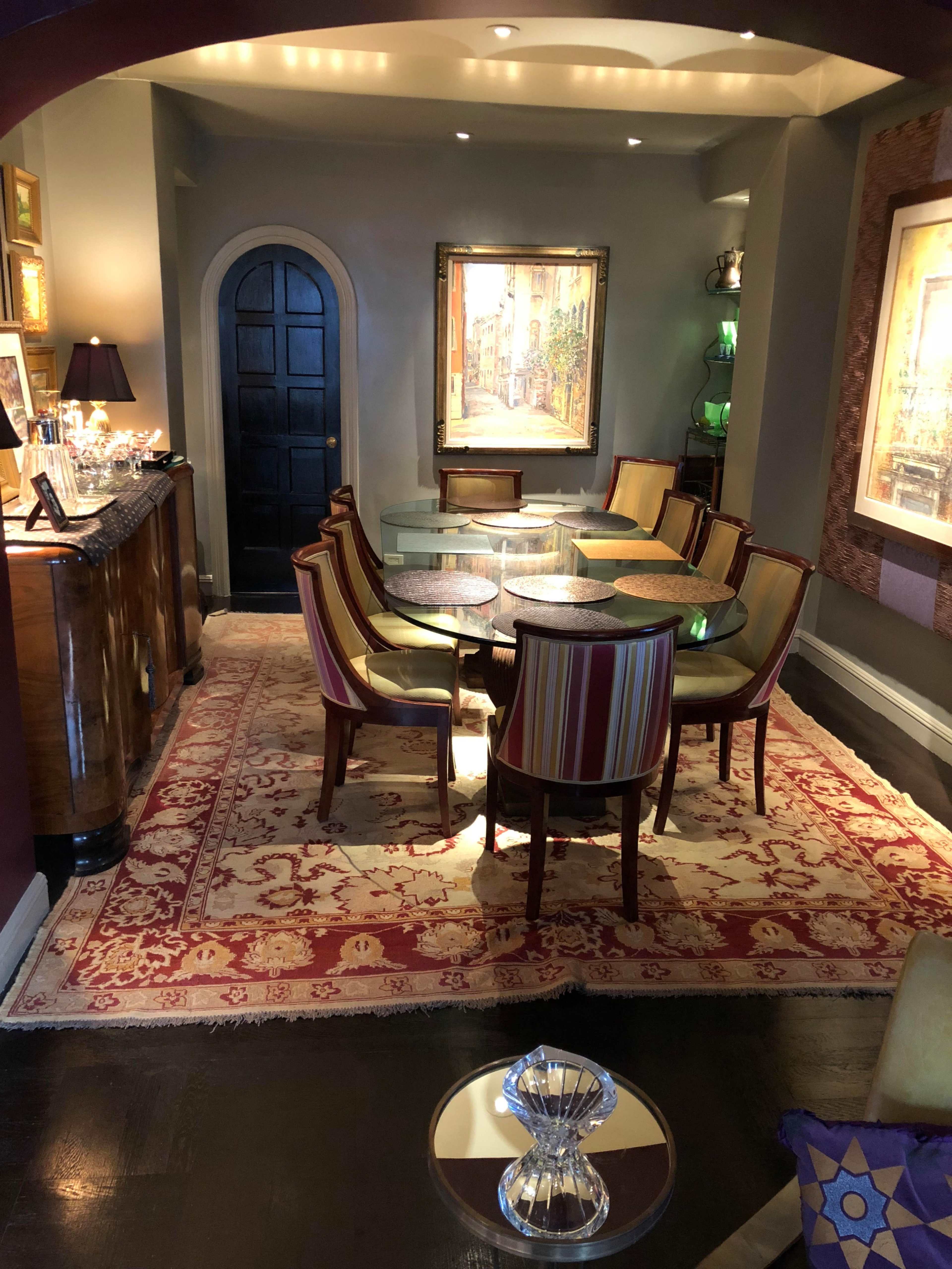 A dining area features a round glass table surrounded by upholstered chairs on a decorative rug, with paintings and a sideboard in the background.