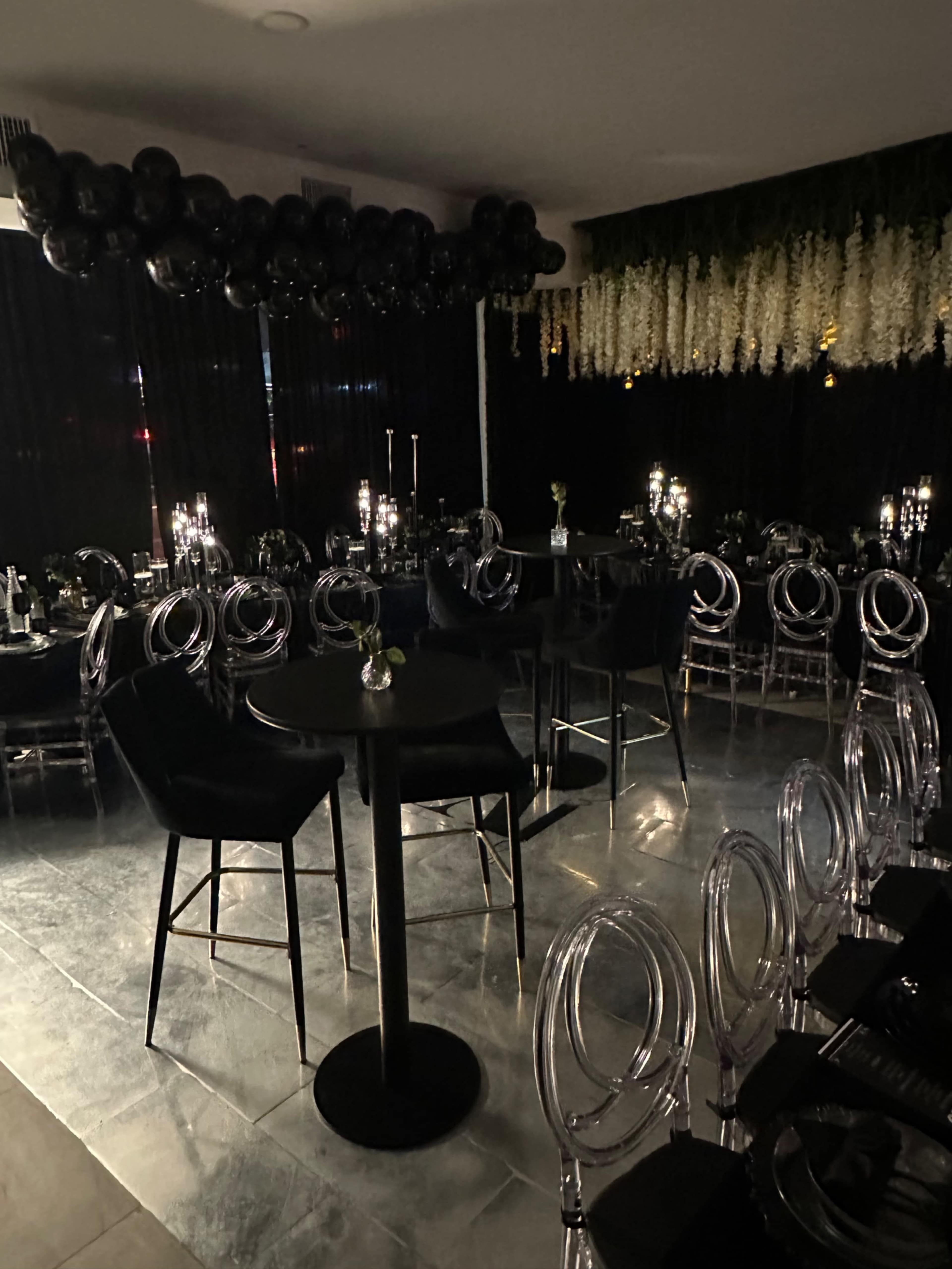A dimly lit event space features black and transparent furniture with a balloon arch and floral decorations overhead.