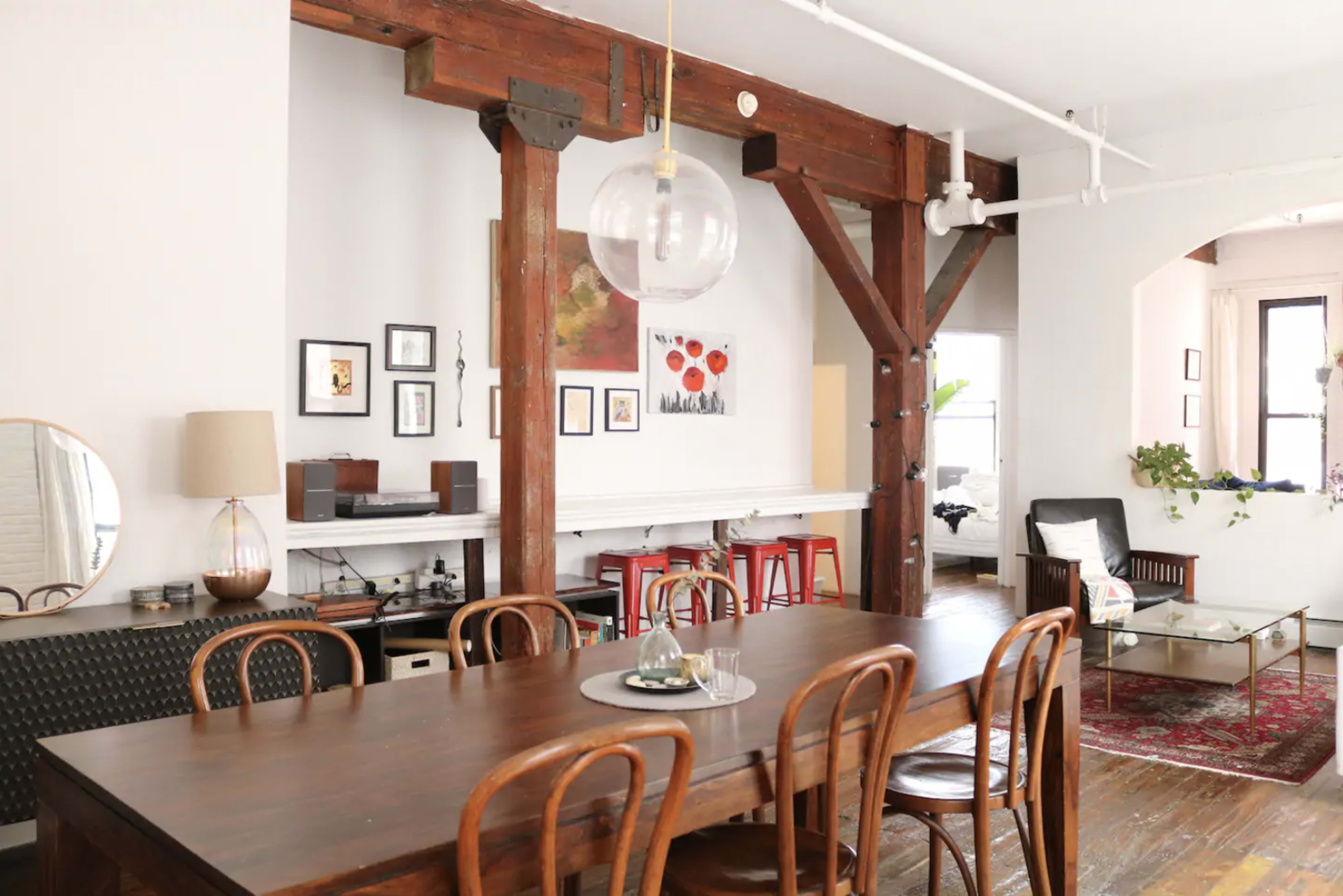 The image shows a spacious dining area featuring a large wooden table surrounded by chairs, with exposed wooden beams and a variety of framed artworks on the walls.