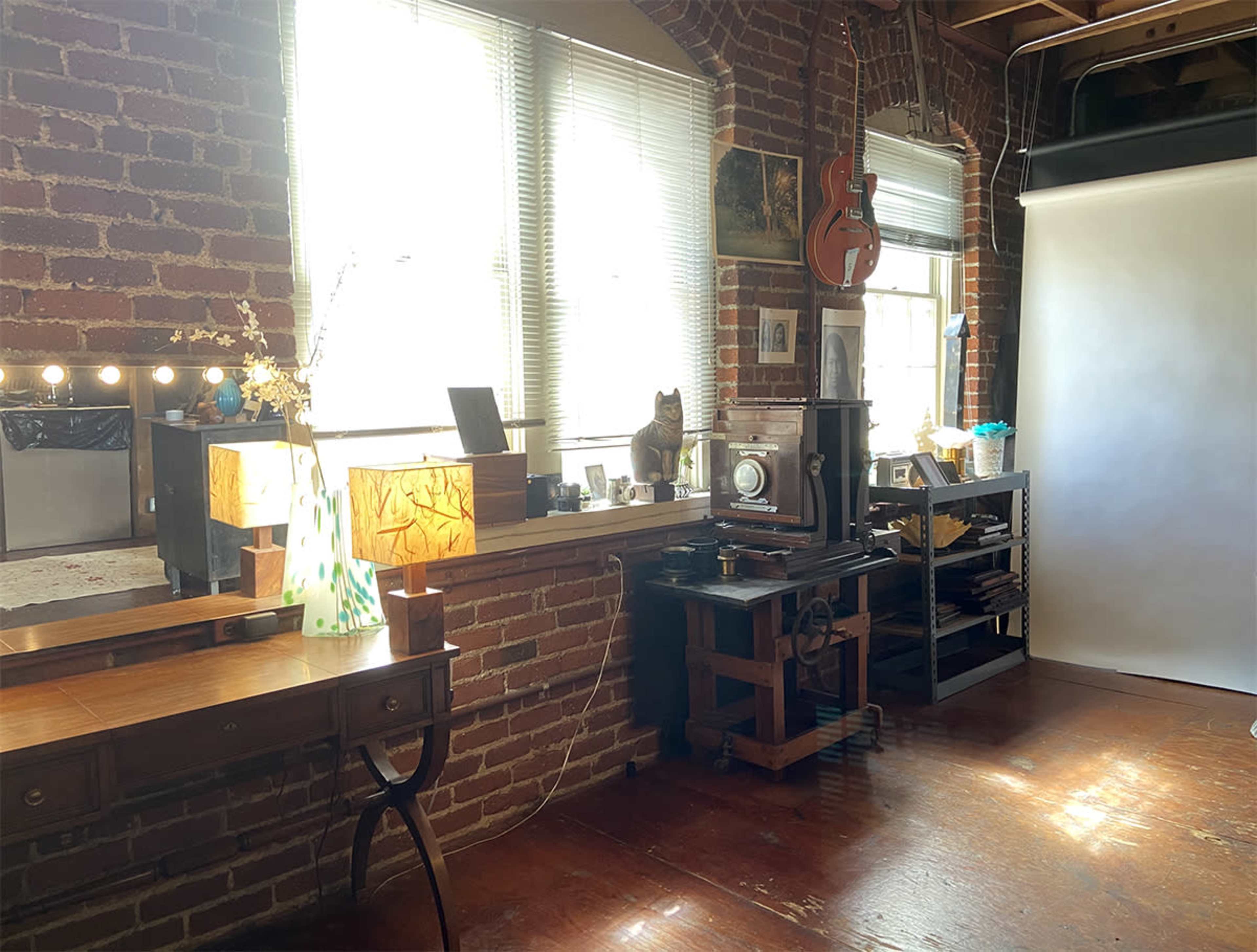 A sunlit room with exposed brick walls, a wooden desk, a vintage camera setup, and various decorative items adorning a long shelf.