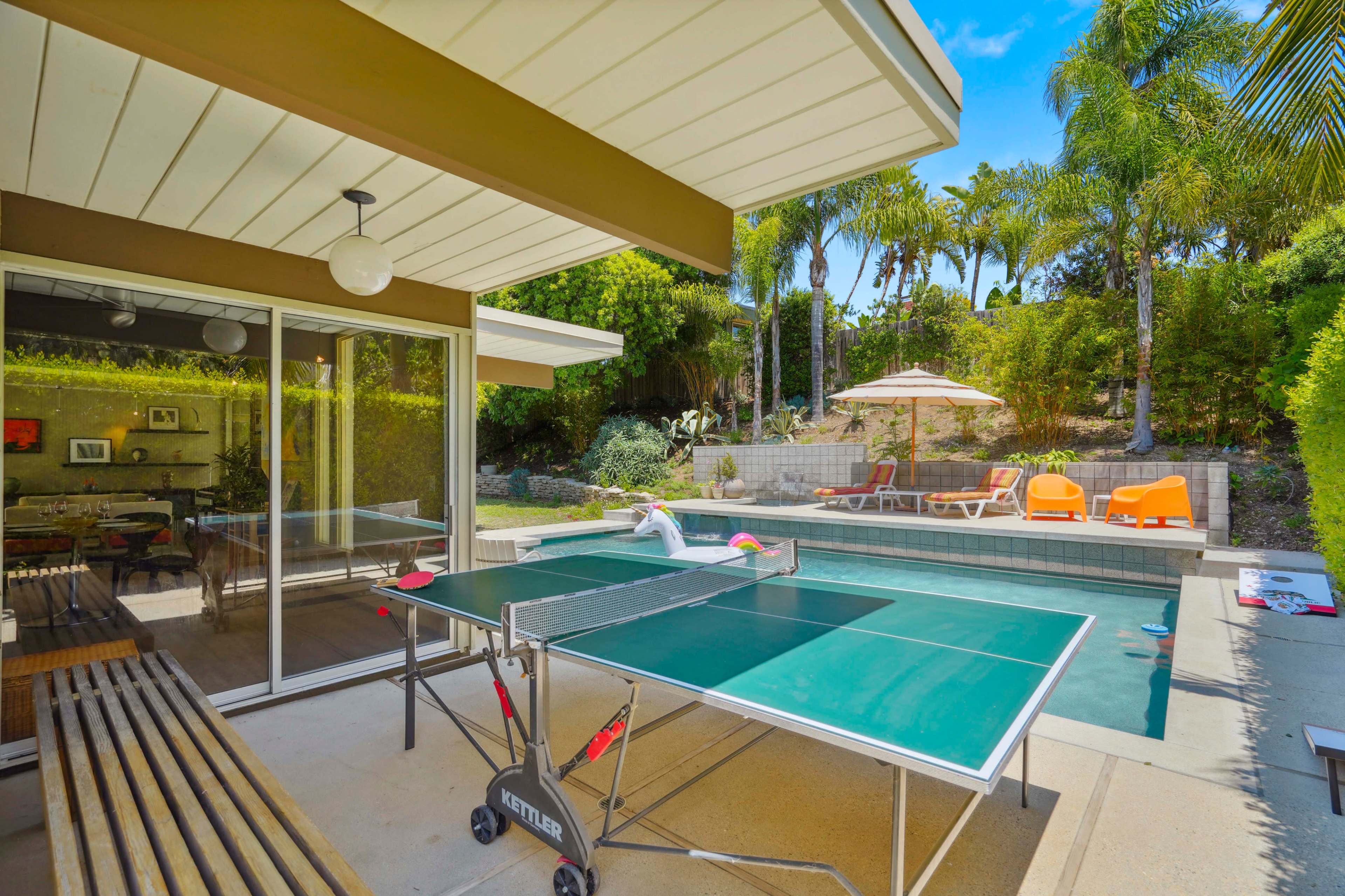 A ping pong table is set up on a patio beside a pool, surrounded by palm trees and lounge chairs.