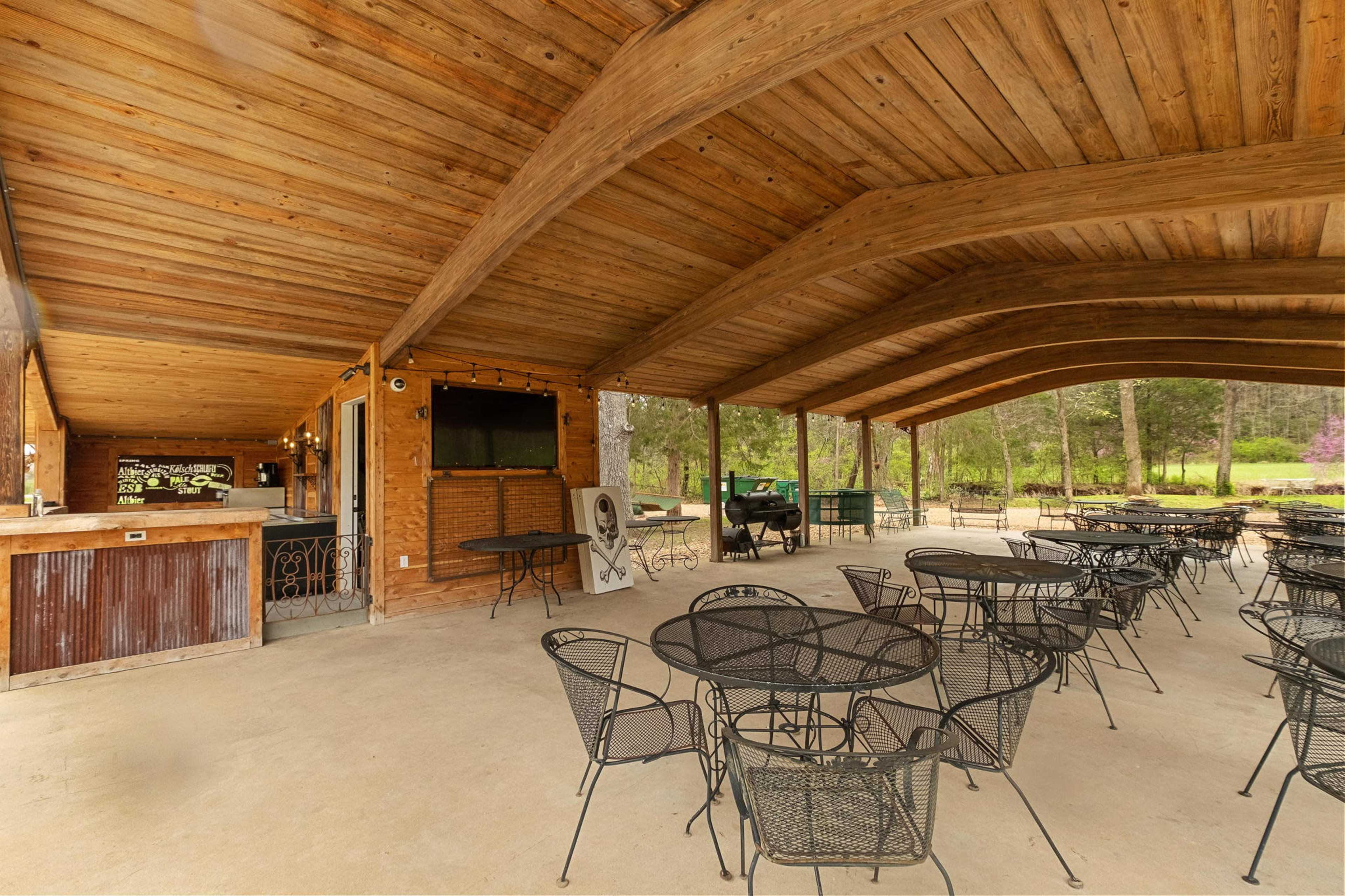 The image shows an outdoor dining area with metal tables and chairs under a large wooden canopy, featuring a bar area in the background.