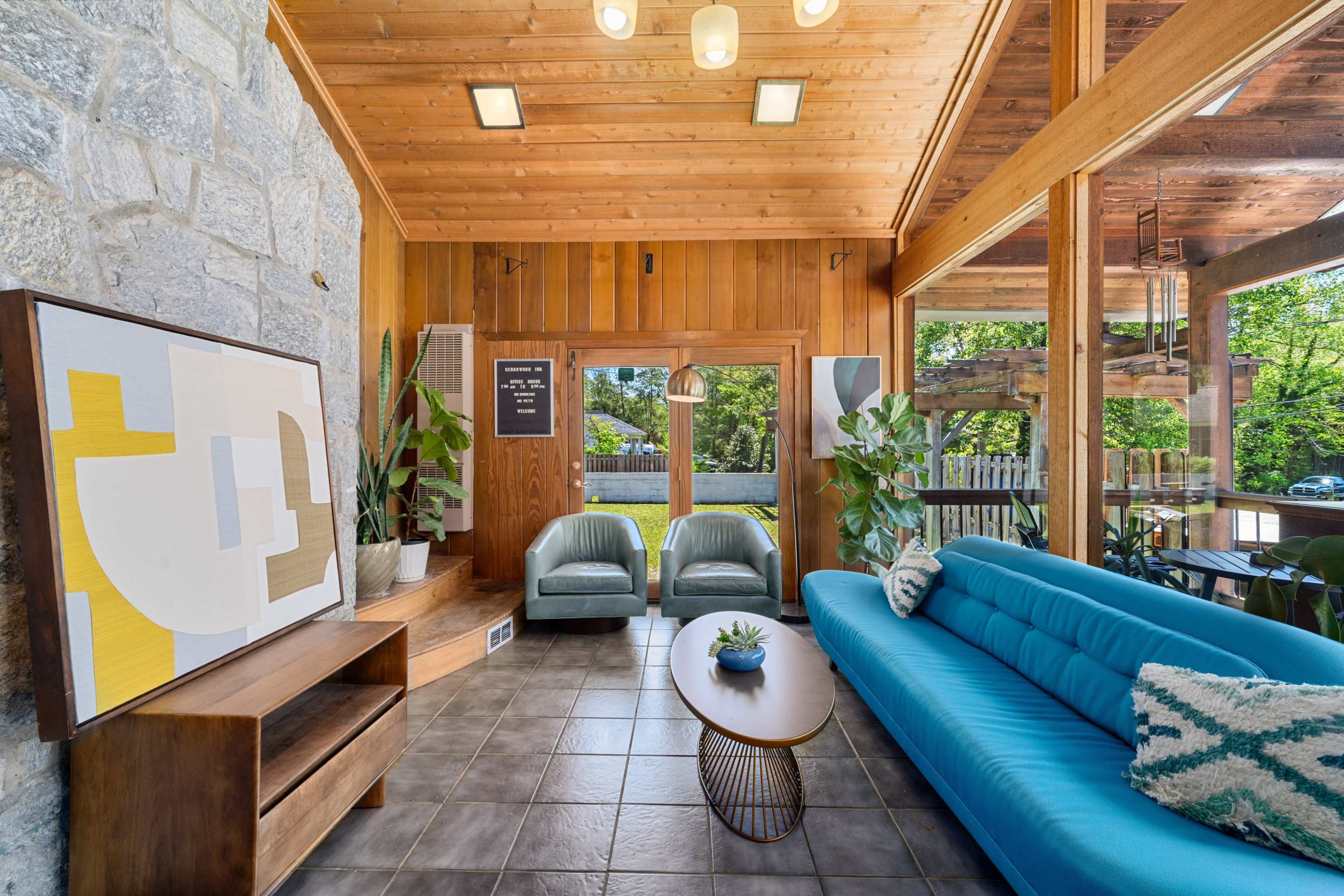 The scene features a modern interior with a blue sofa, two green chairs, a wooden coffee table, and large windows surrounded by plants and stone walls.