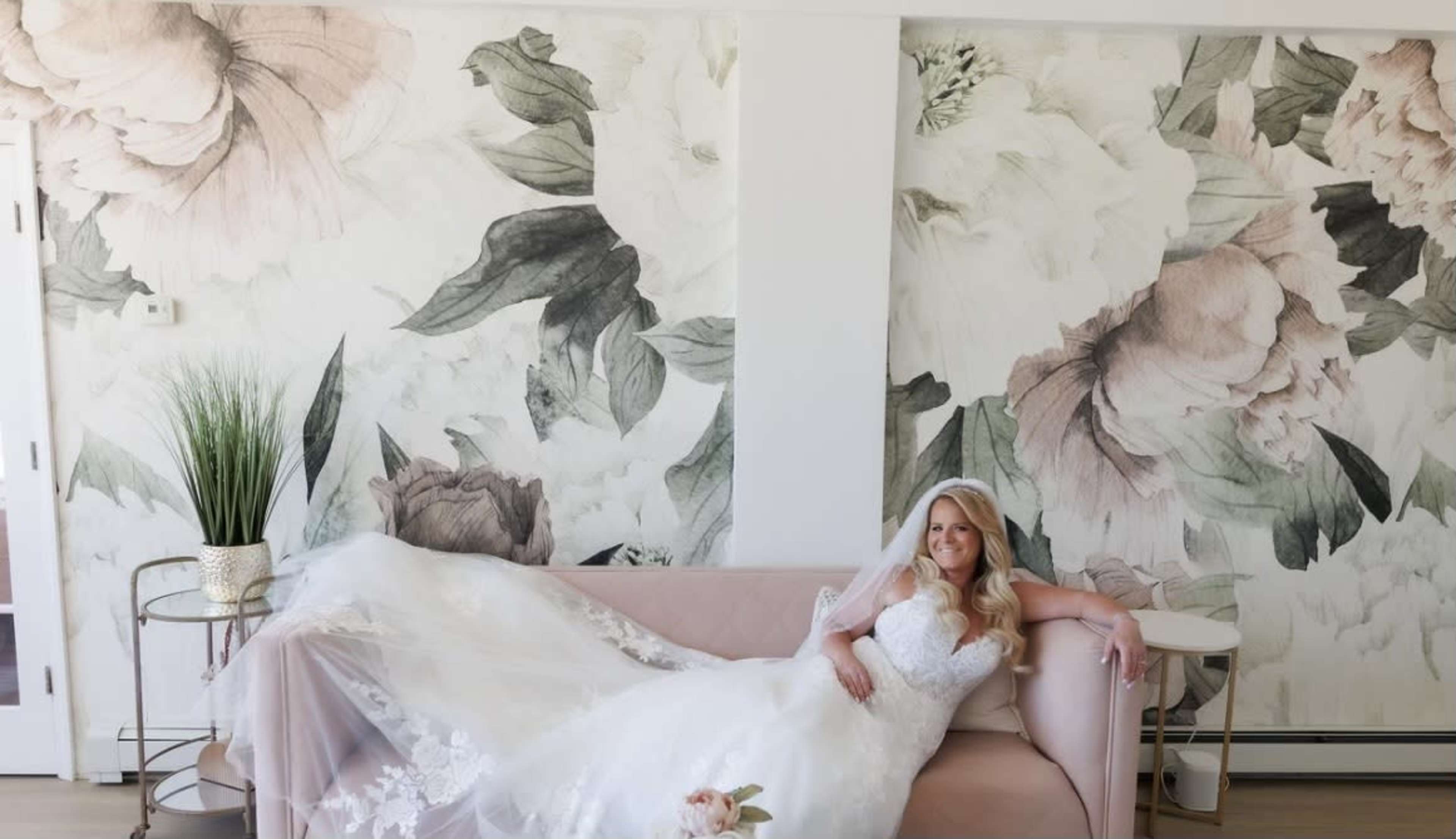 A bride in a wedding dress lounges on a pink couch in front of floral wall murals.
