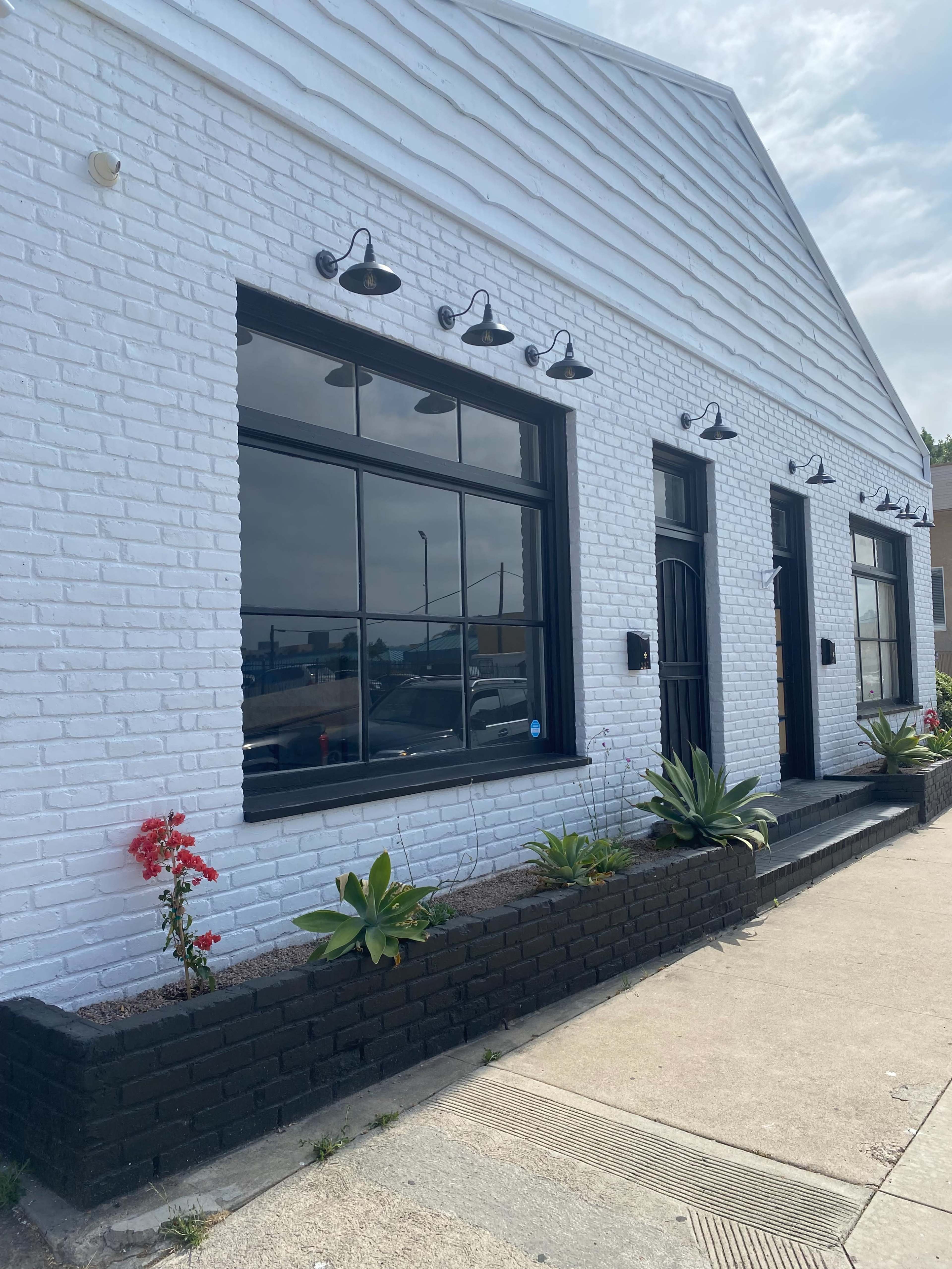 The image shows a white brick building featuring large windows, modern outdoor lighting fixtures, and planters with green plants and red flowers along the walkway.