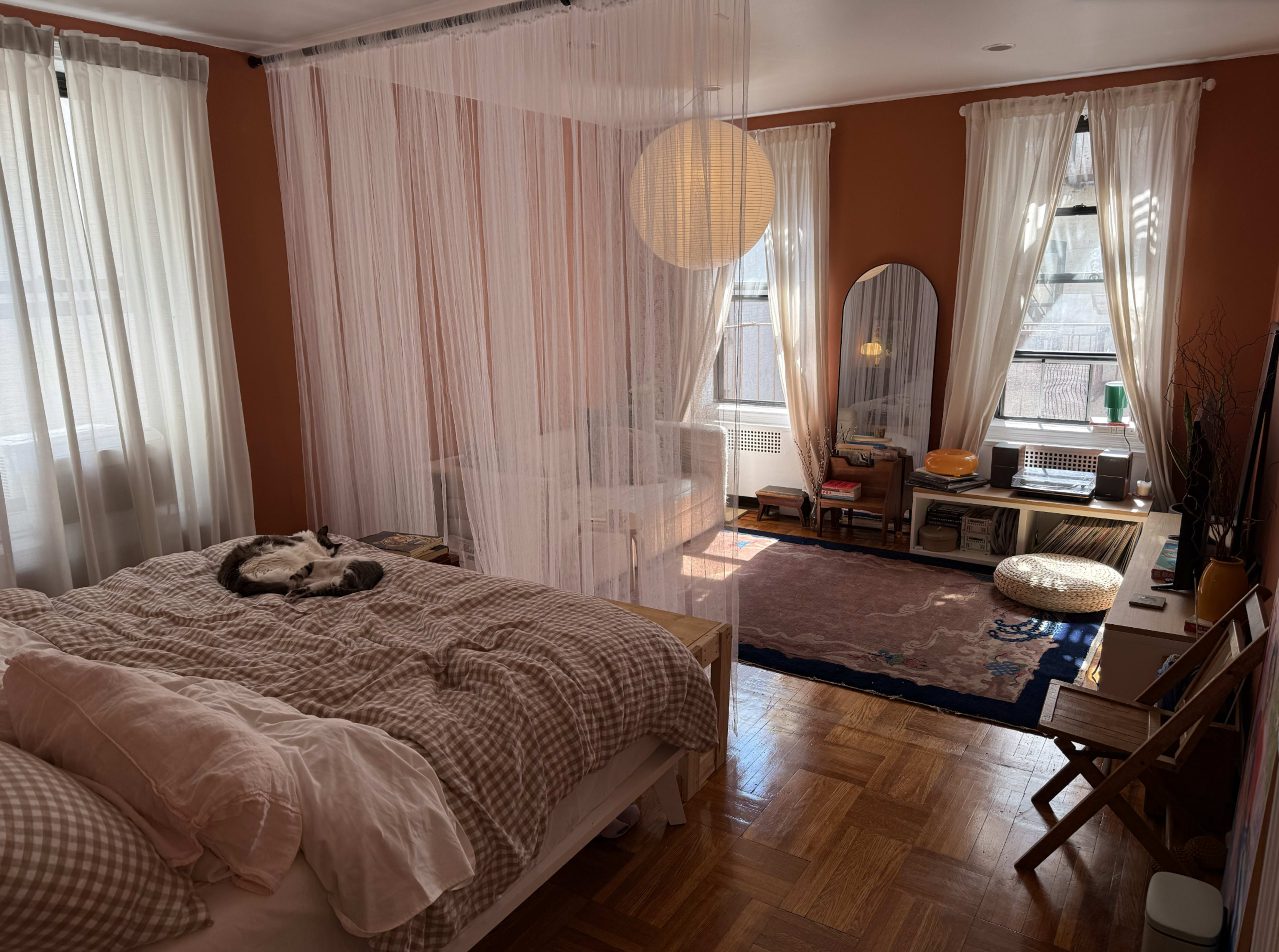 The image shows a cozy bedroom featuring a bed with a patterned blanket, a cat resting on it, sheer curtains, and a sitting area with bookshelves and an armchair.