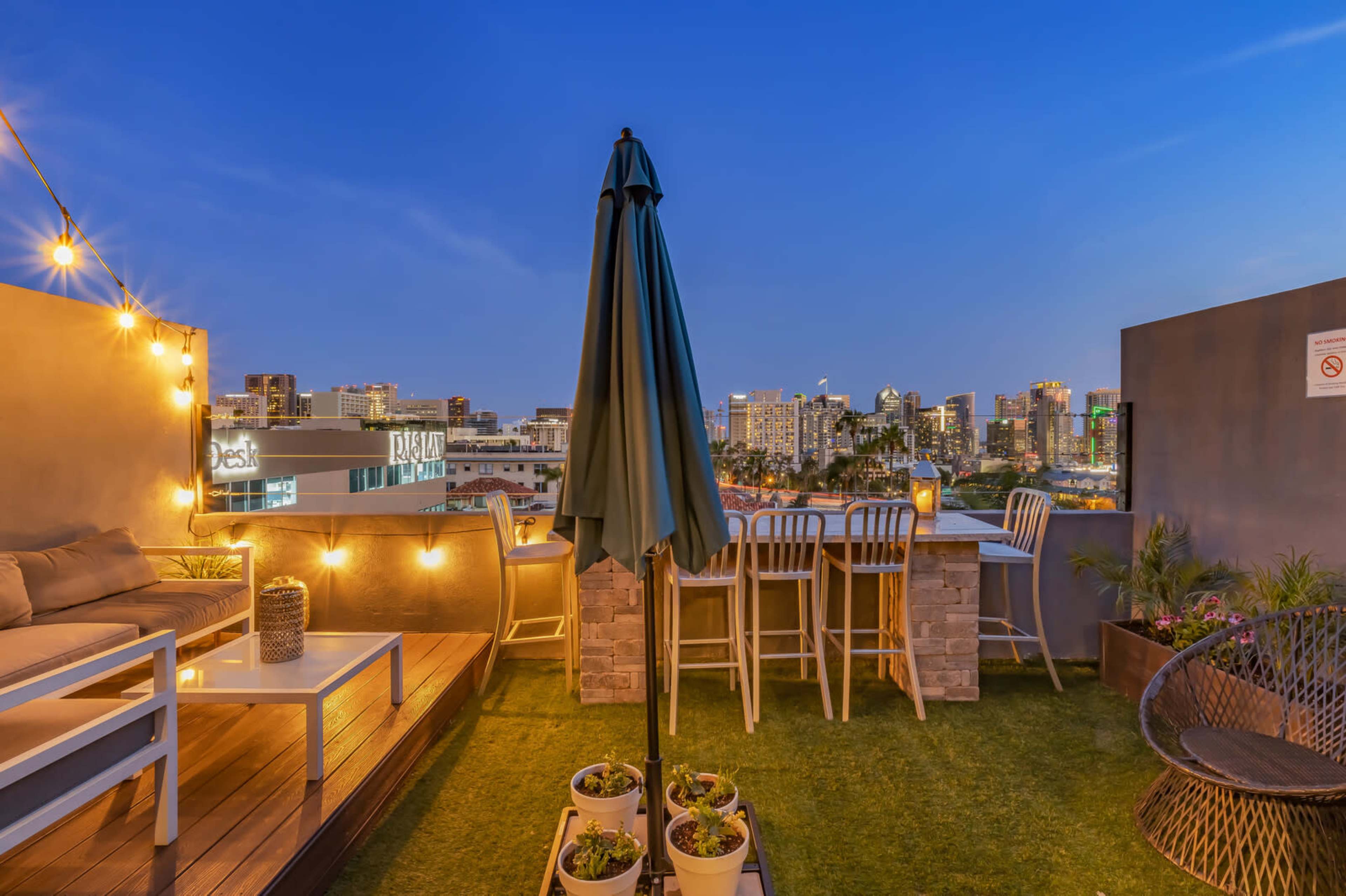 A rooftop patio features a table with tall chairs, a shaded umbrella, and potted plants, overlooking a city skyline at dusk.