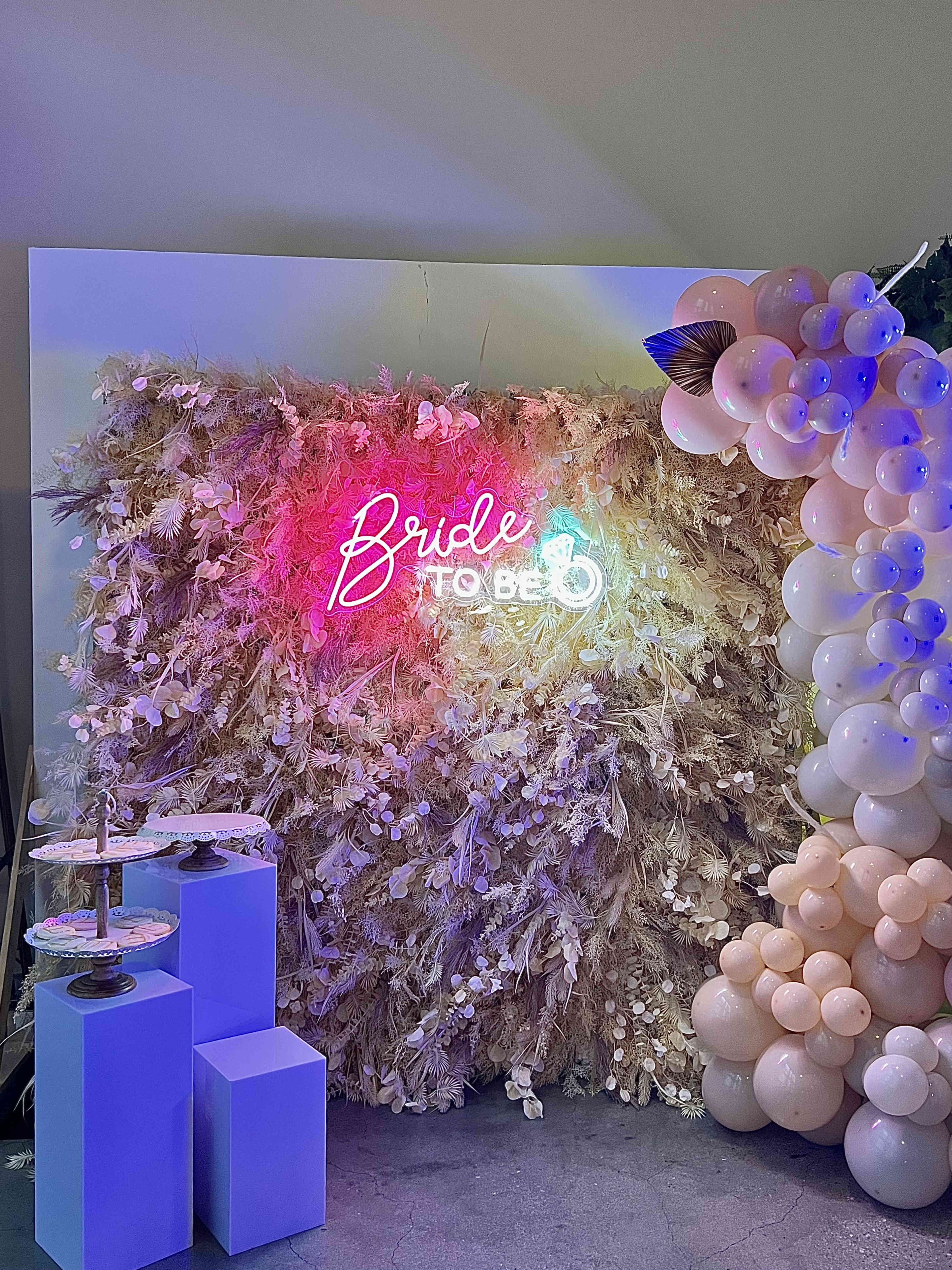 A decorative backdrop with a vibrant "Bride to Be" neon sign surrounded by dried flowers and an artistic arrangement of pastel-colored balloons.