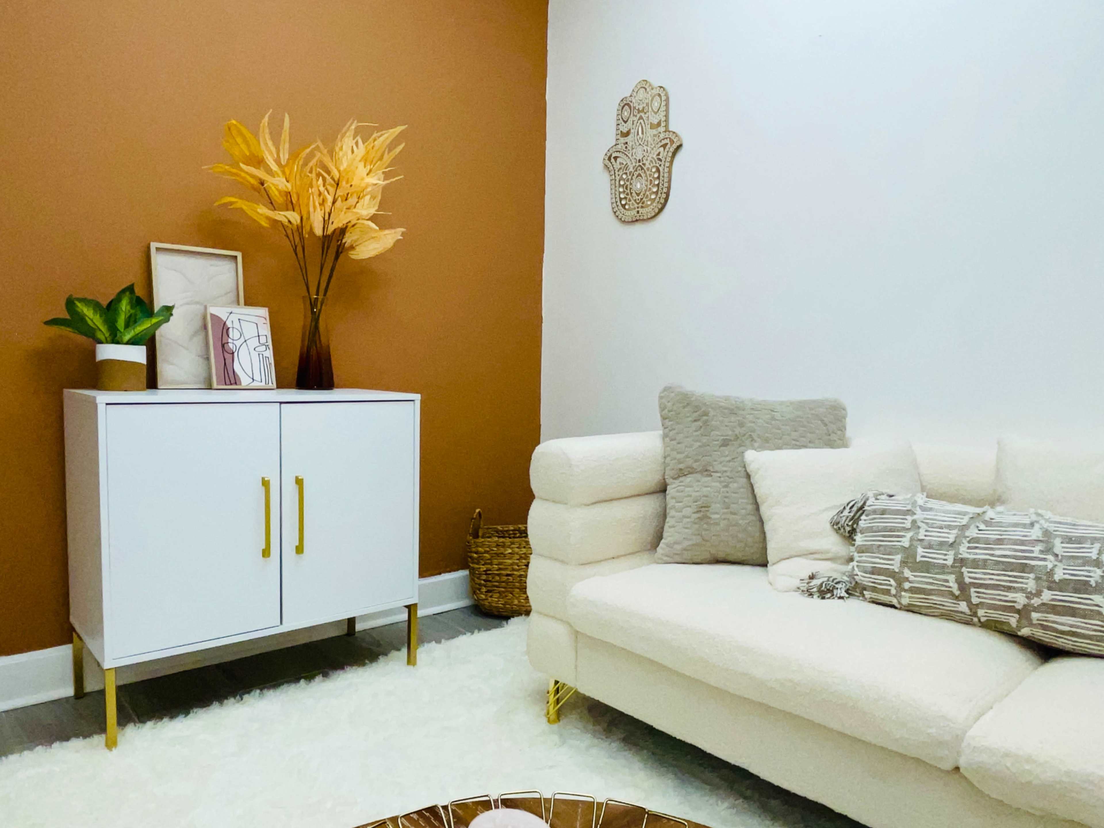 A cozy living room with a white sofa, a decorative cabinet, and a brown accent wall, complemented by plants and warm decor.