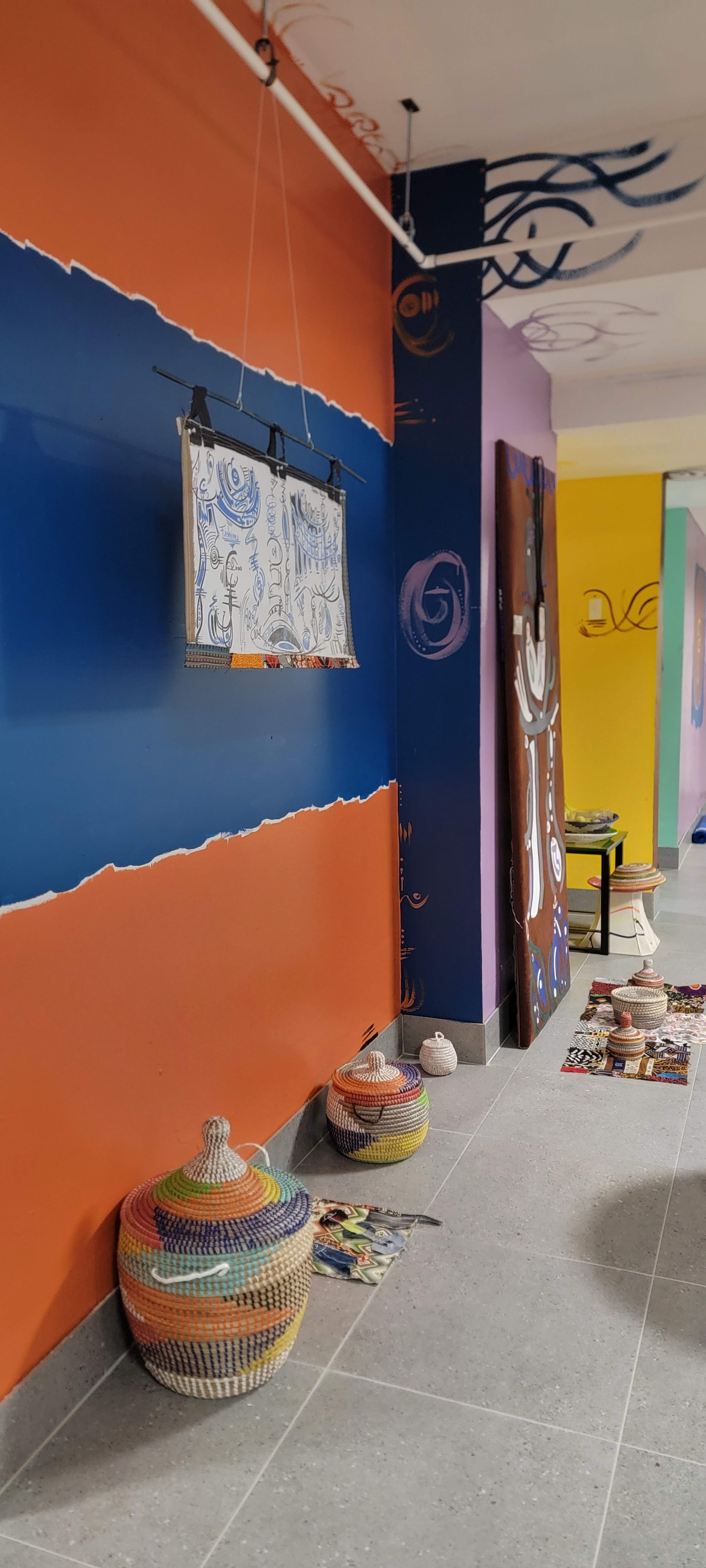 The image shows a colorful interior wall with orange, purple, and blue sections, featuring woven baskets on the floor and a framed artwork hanging above.