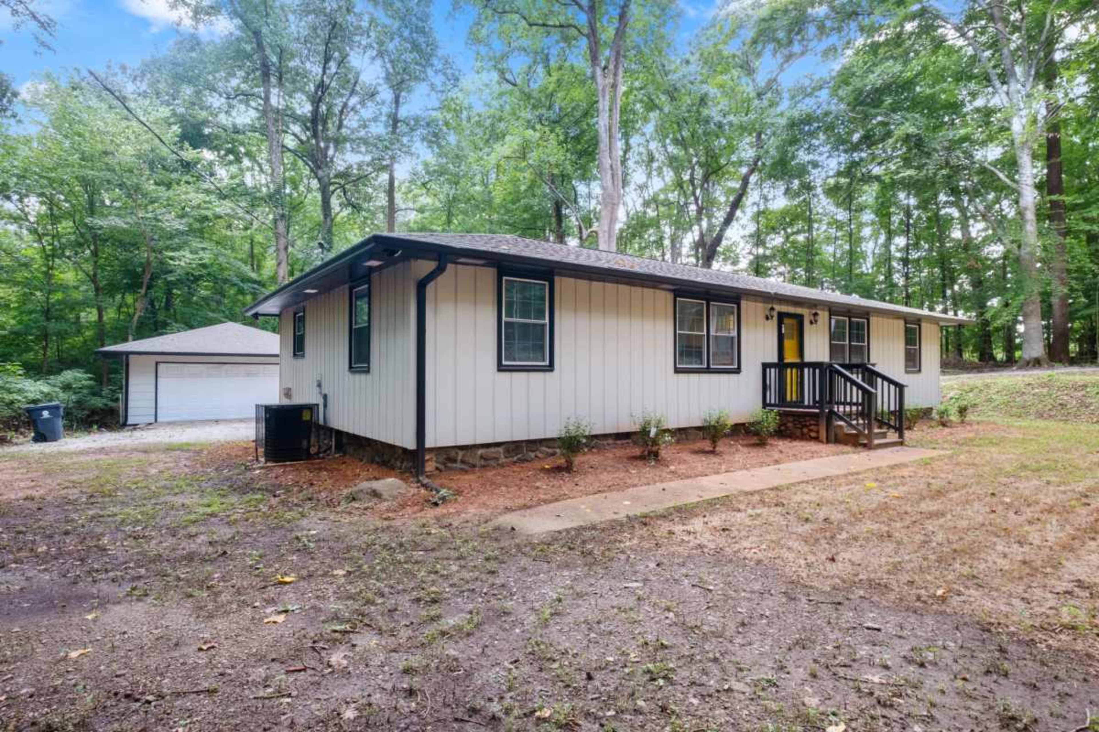 A single-story house with a light-colored exterior and a dark roof is situated next to a small garage in a wooded area.