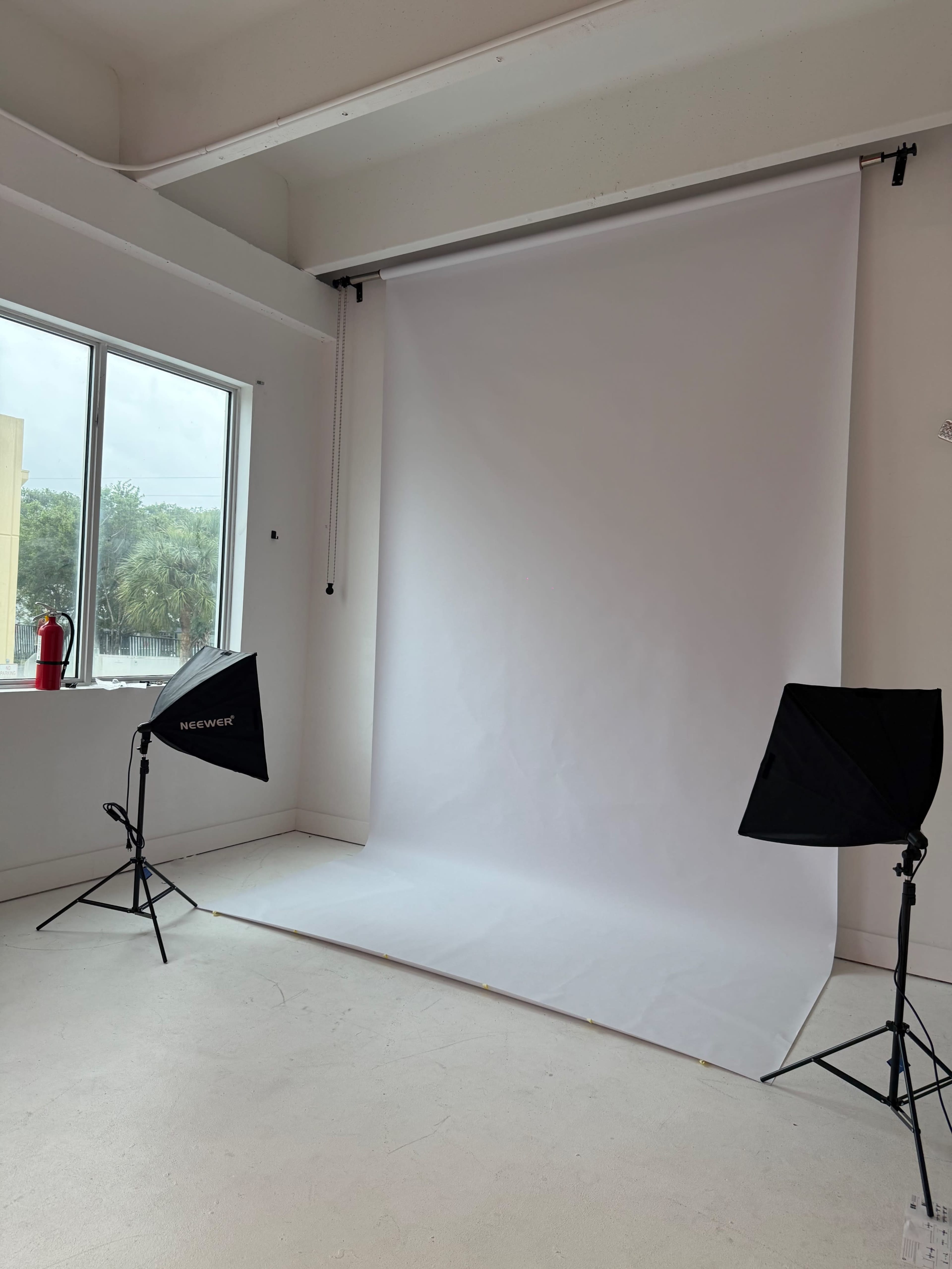 The image shows a photography studio with a white backdrop, two softbox lights positioned on either side, and a window in the background.