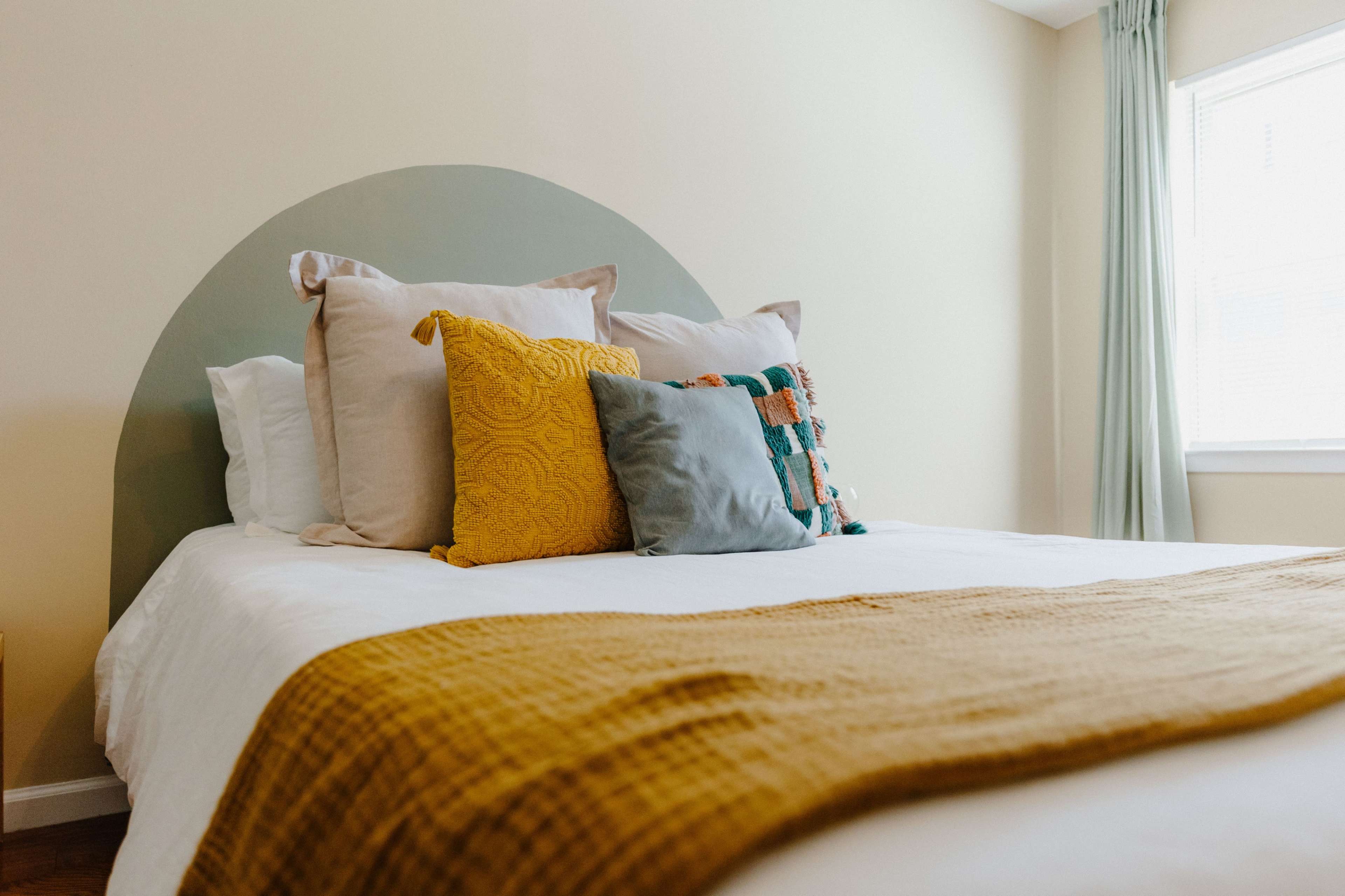 A neatly made bed with a variety of pillows and a textured yellow blanket is positioned against a light-colored wall in a well-lit room.