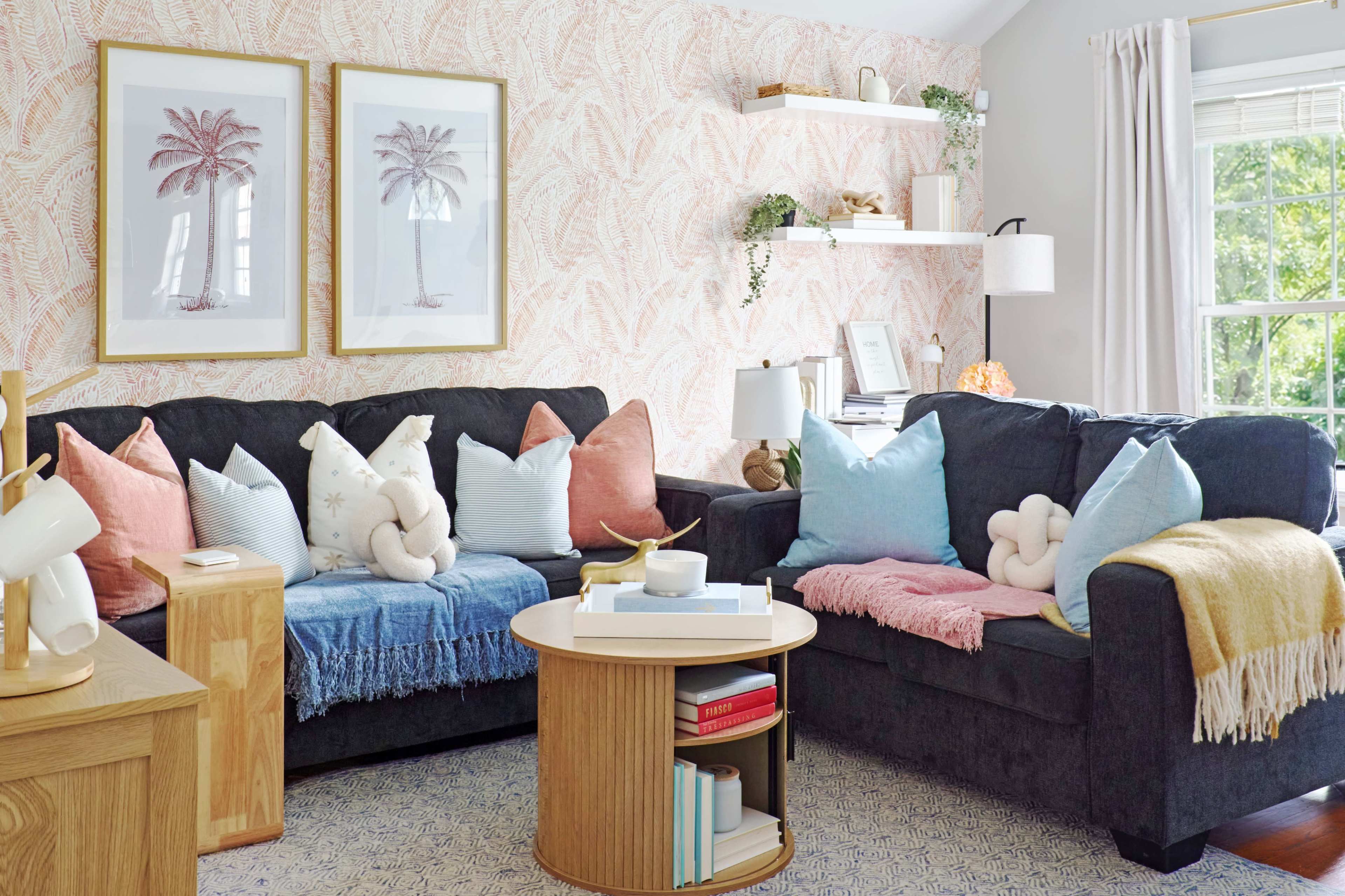 The living room features two dark couches adorned with colorful cushions, a round coffee table in the center, and framed palm tree artwork on the wall.