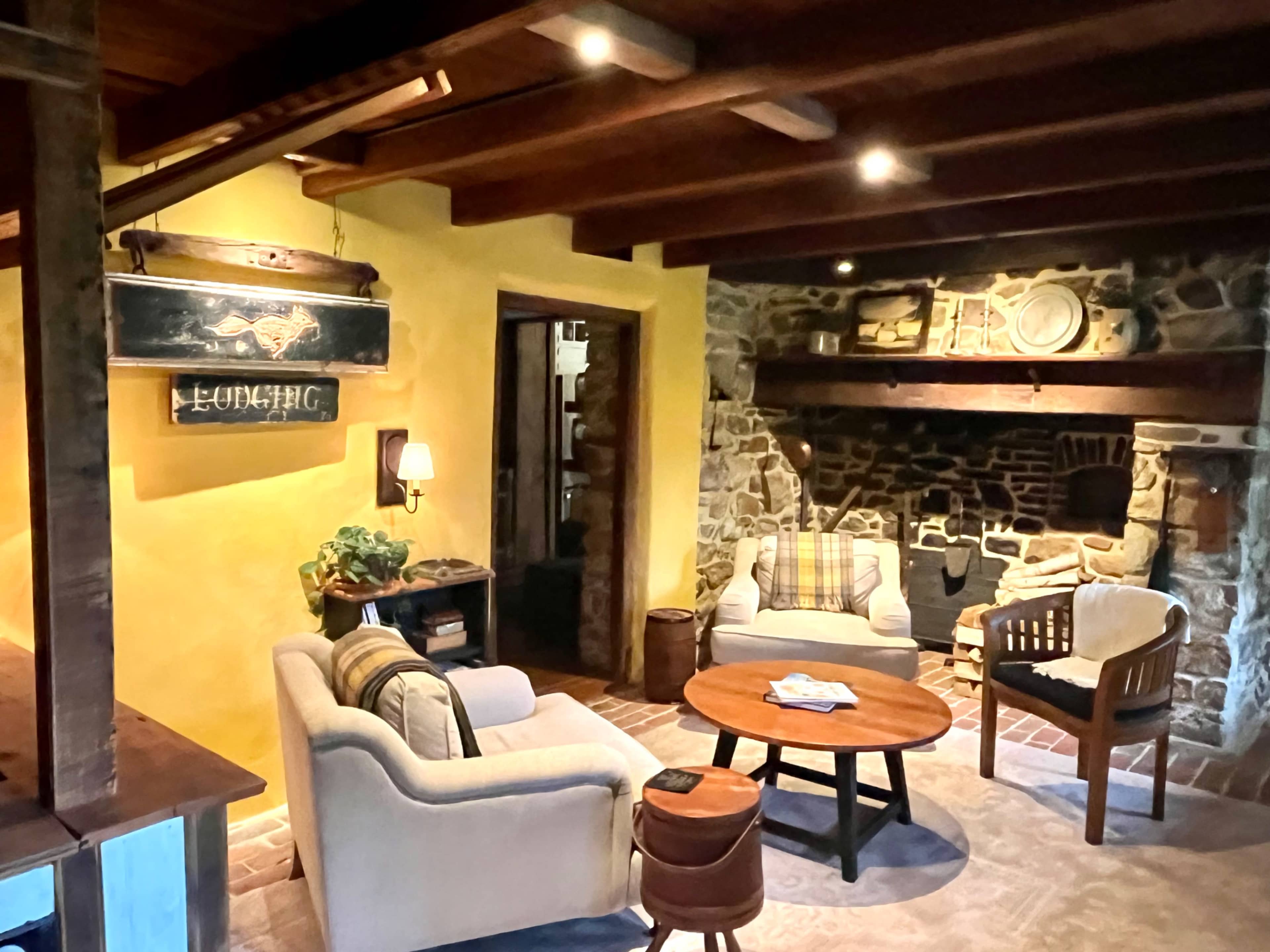 A cozy living room features a stone fireplace, wooden beams, and comfortable seating arranged around a round table.