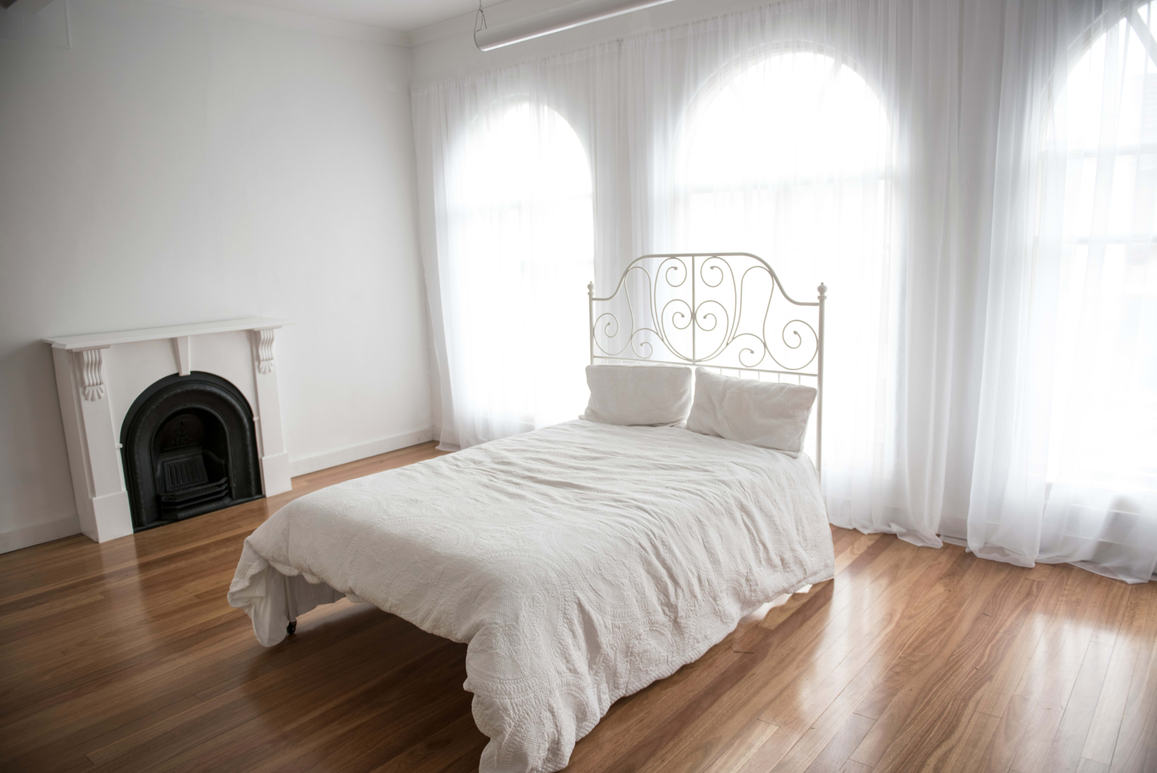 The image shows a simple bedroom featuring a white bed with a decorative headboard and large windows covered by sheer curtains.