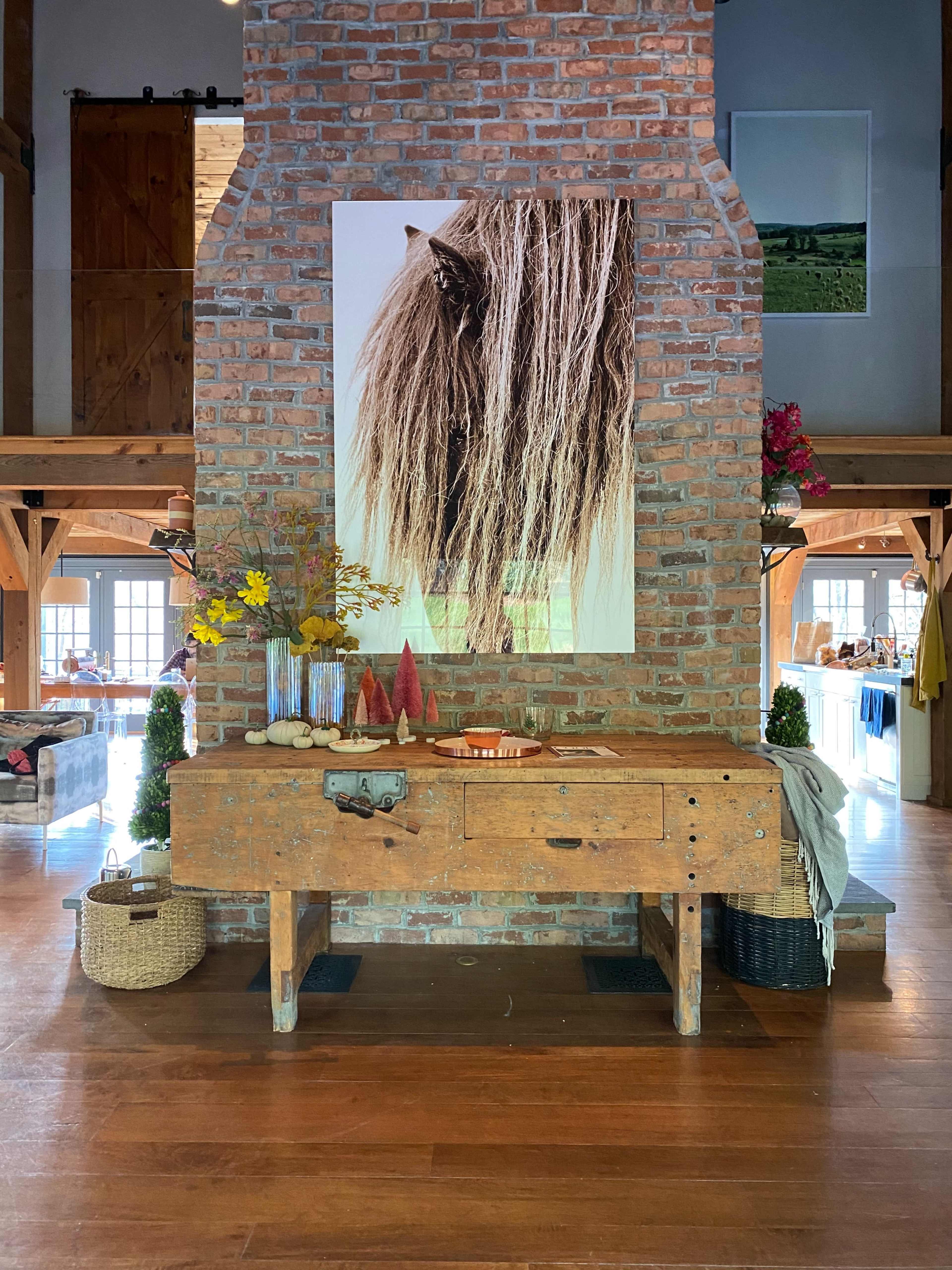 A rustic wooden table with decorative items and flowers is positioned in front of a large brick column featuring a large photo of a horse's mane.
