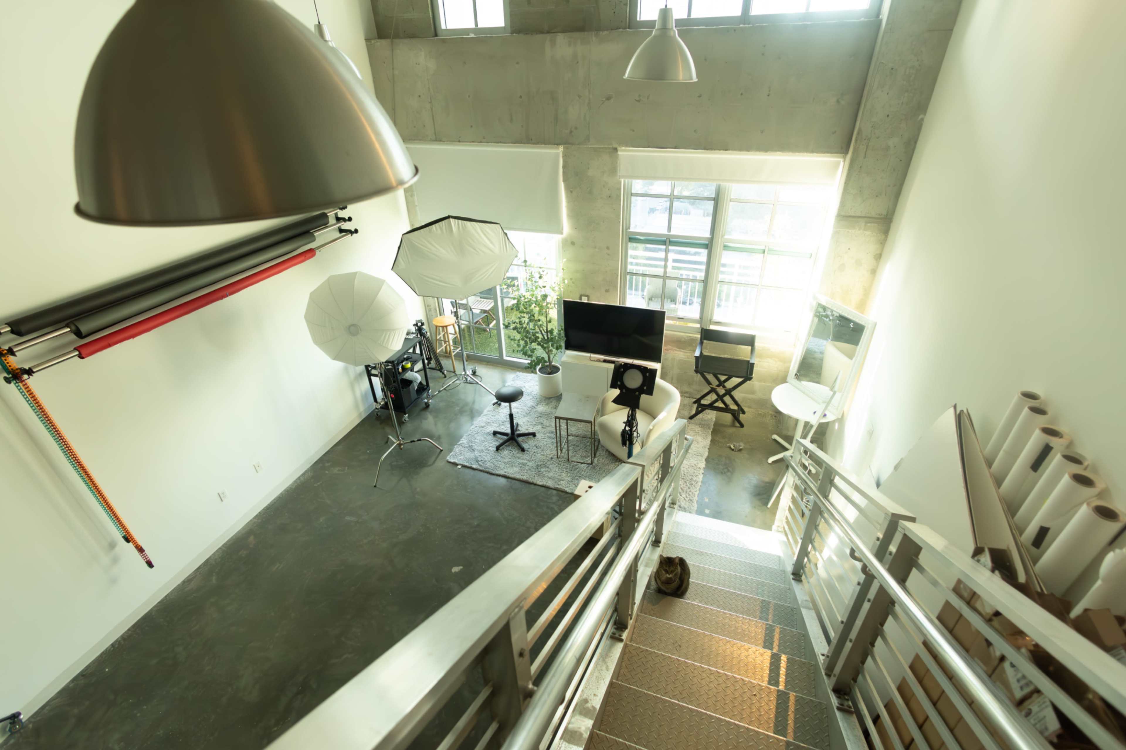 The image features a spacious studio with concrete walls, several softbox lights, a computer setup, and large windows allowing natural light to enter.