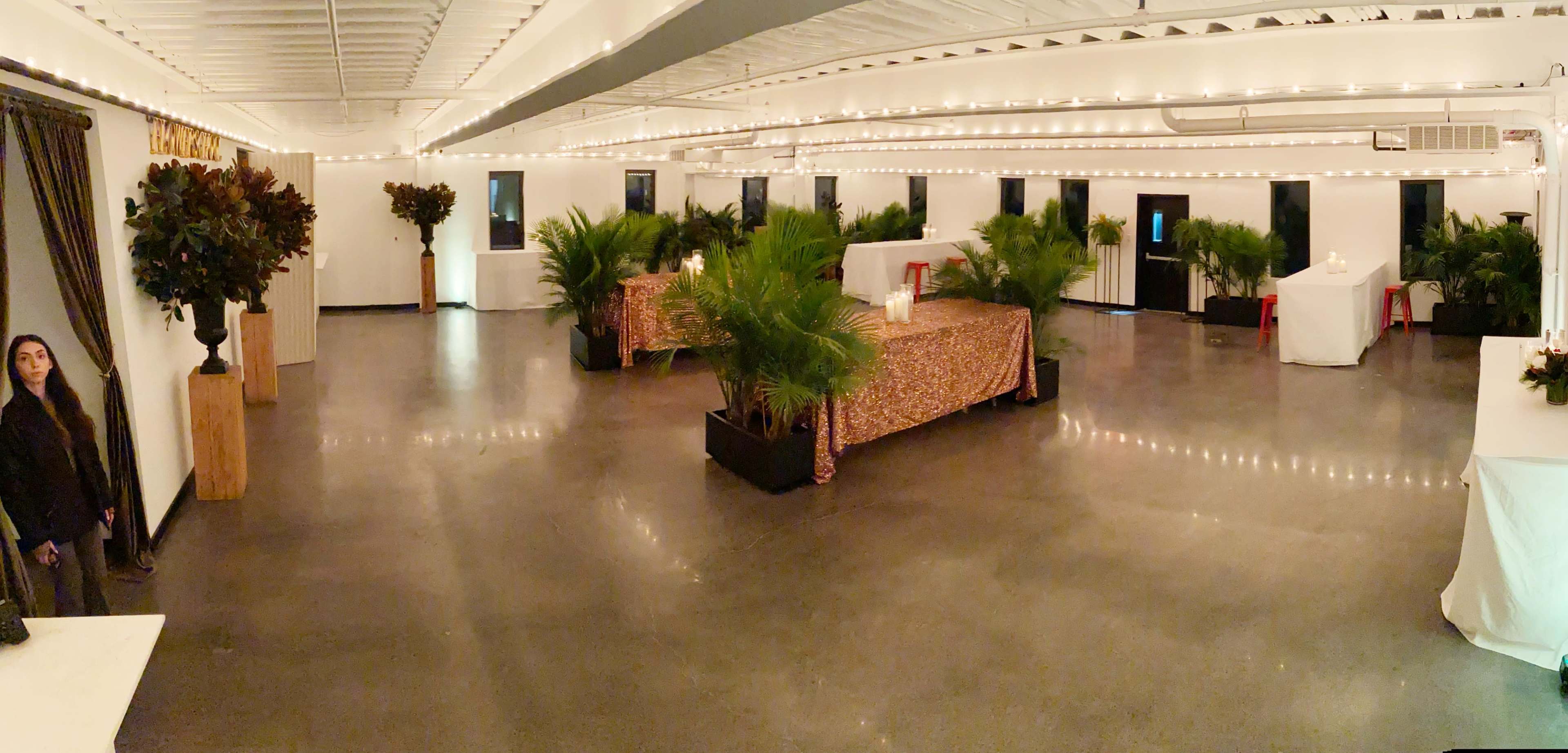 A spacious, well-lit room decorated with potted palm plants and tables covered with tablecloths, ready for an event.