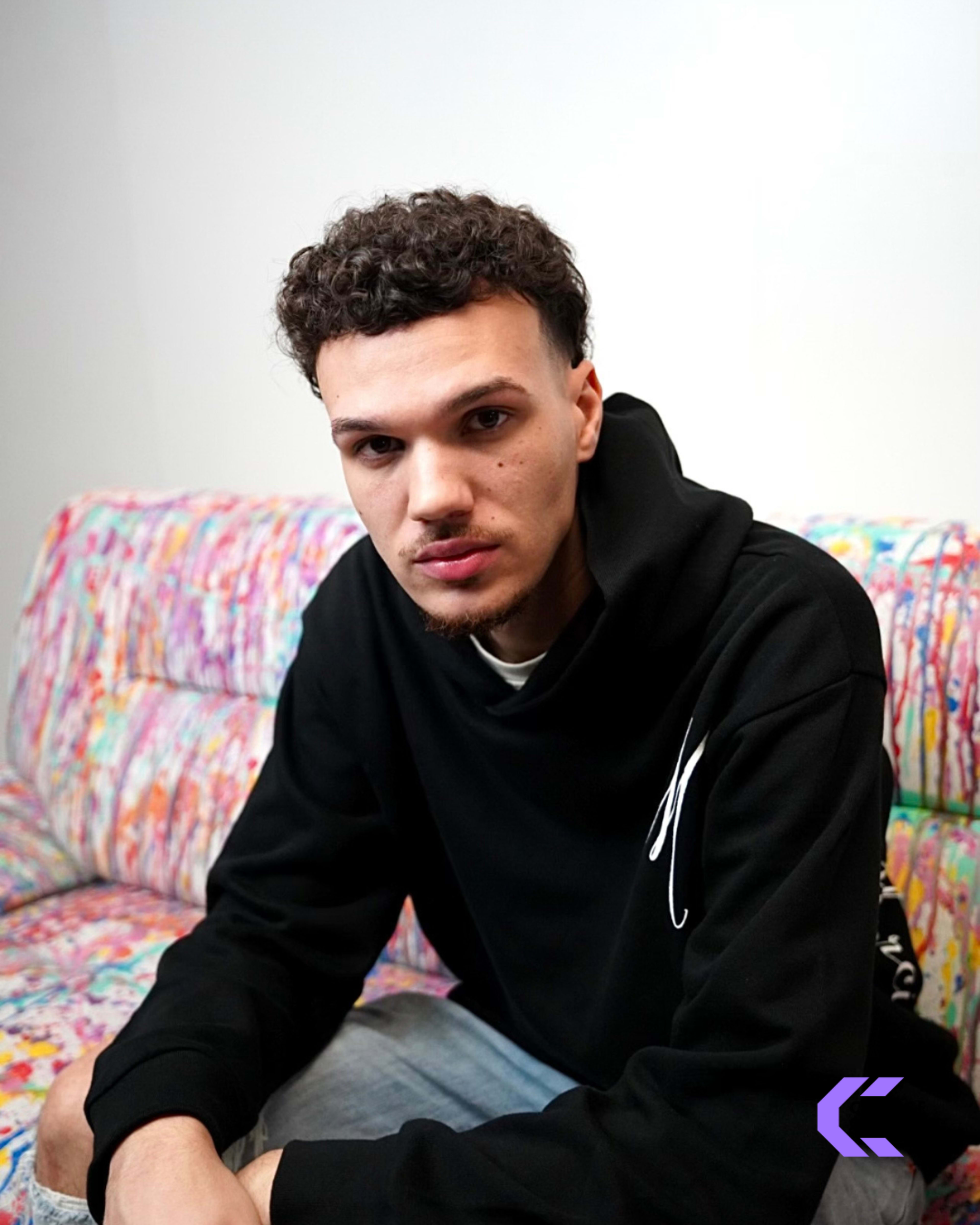 A man with curly hair is sitting on a colorful, patterned couch while wearing a black hoodie.