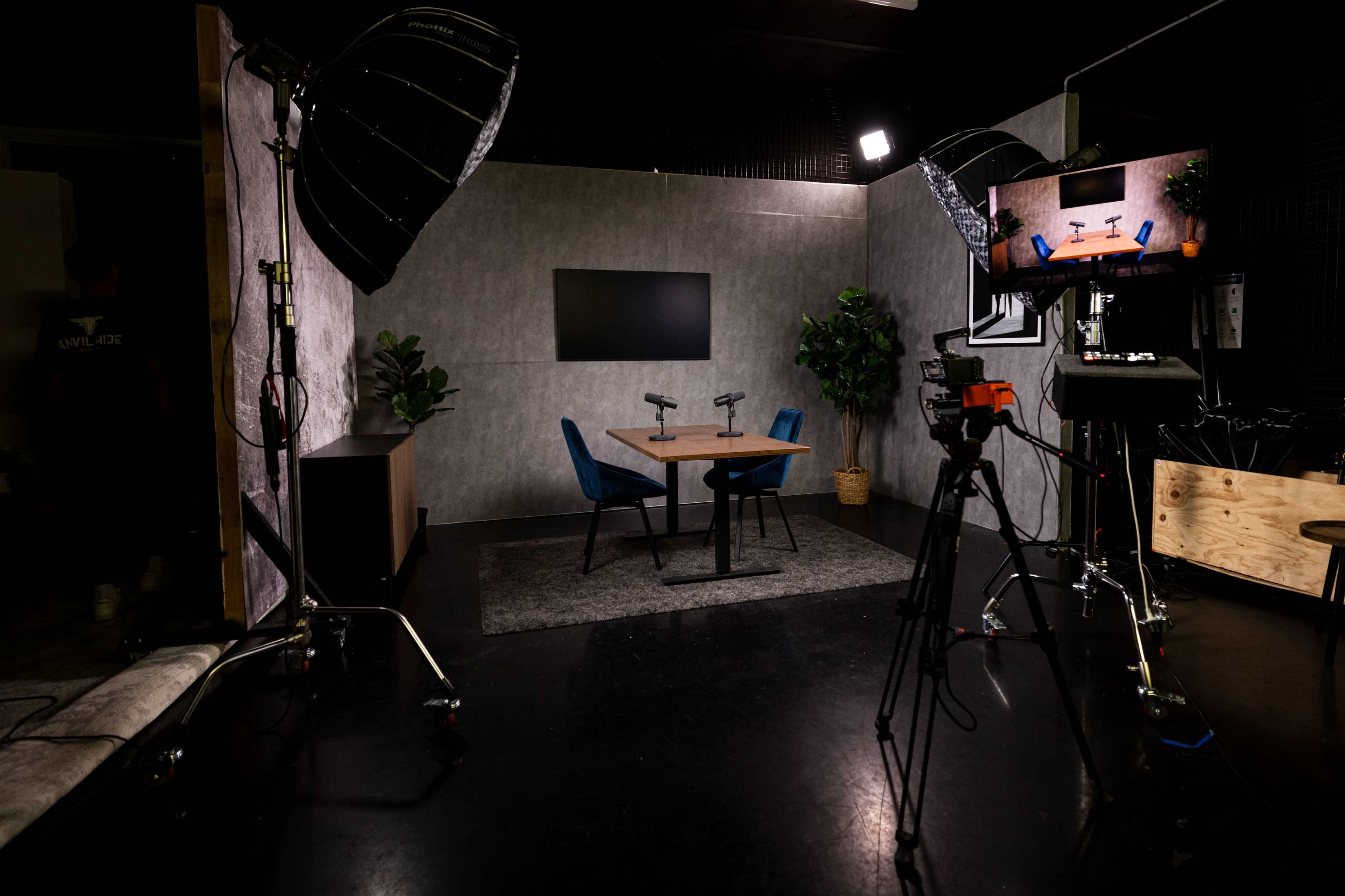 The image shows a modern studio setup with a table, two blue chairs, and cameras positioned for filming, surrounded by lighting equipment and greenery.